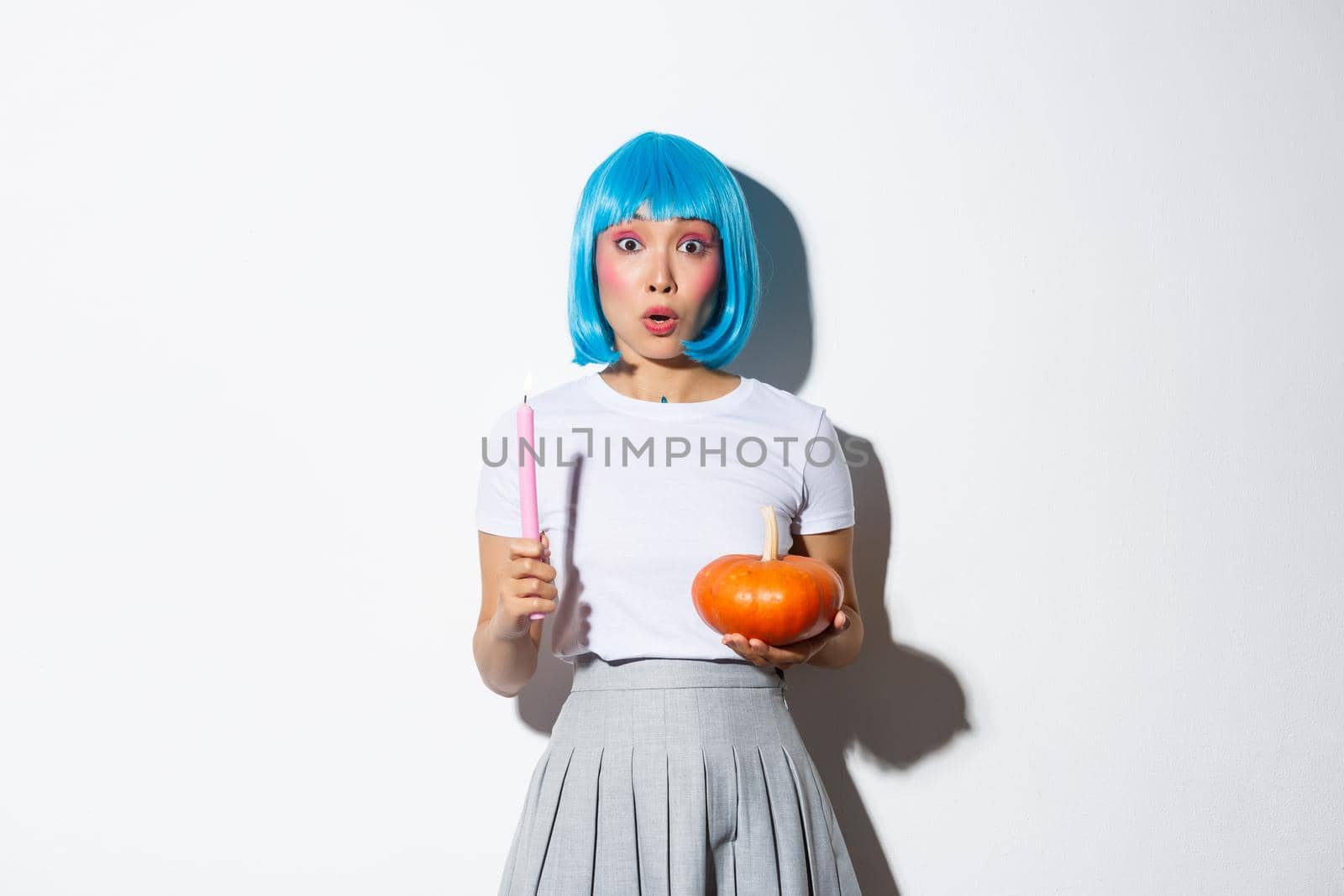 Concept of halloween. Portrait of surprised asian female holding pumpkin and candle, wearing blue wig, gasping amazed as standing over white background.
