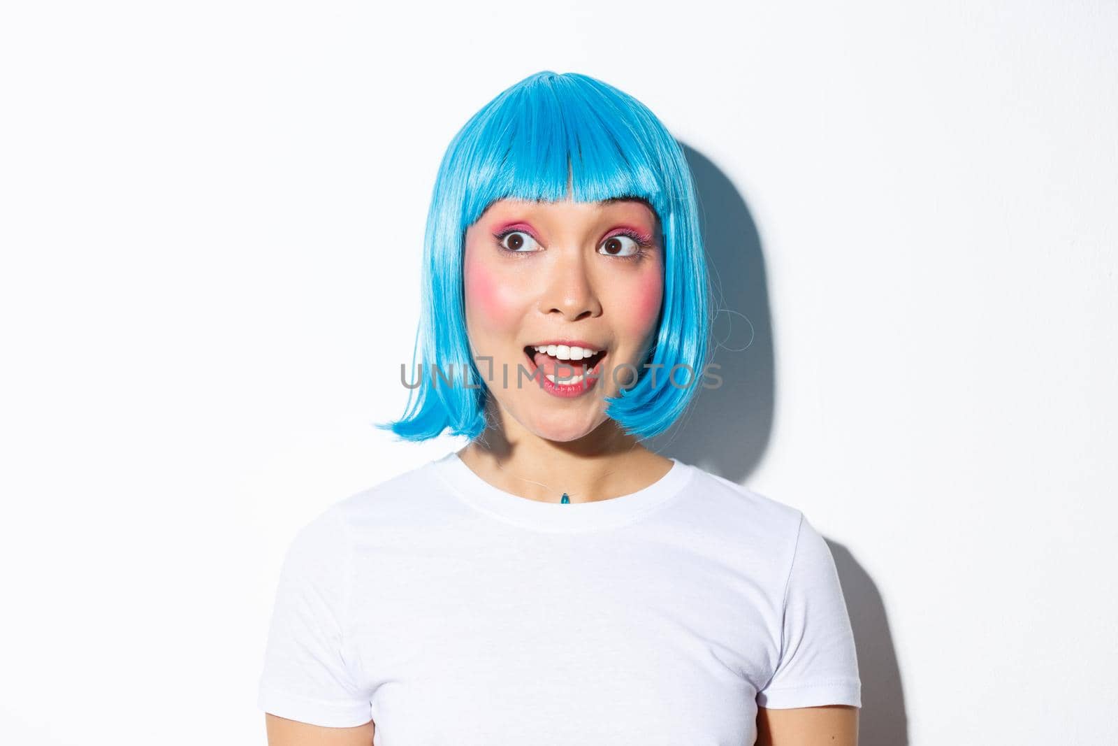 Close-up of surprised cute asian woman in blue wig, looking at halloween banner, standing over white background.