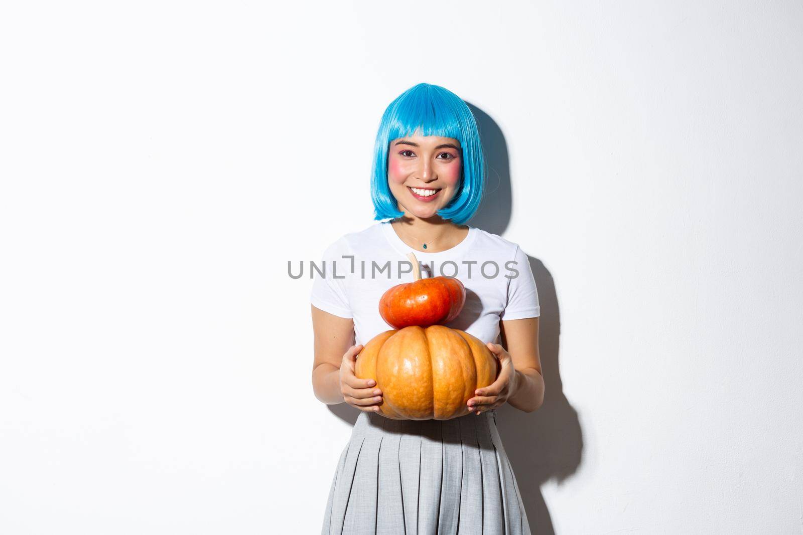 Adorable asian woman in blue wig holding two cute pumpkins and smiling at camera, wearing schoolgirl outfit for halloween party by Benzoix