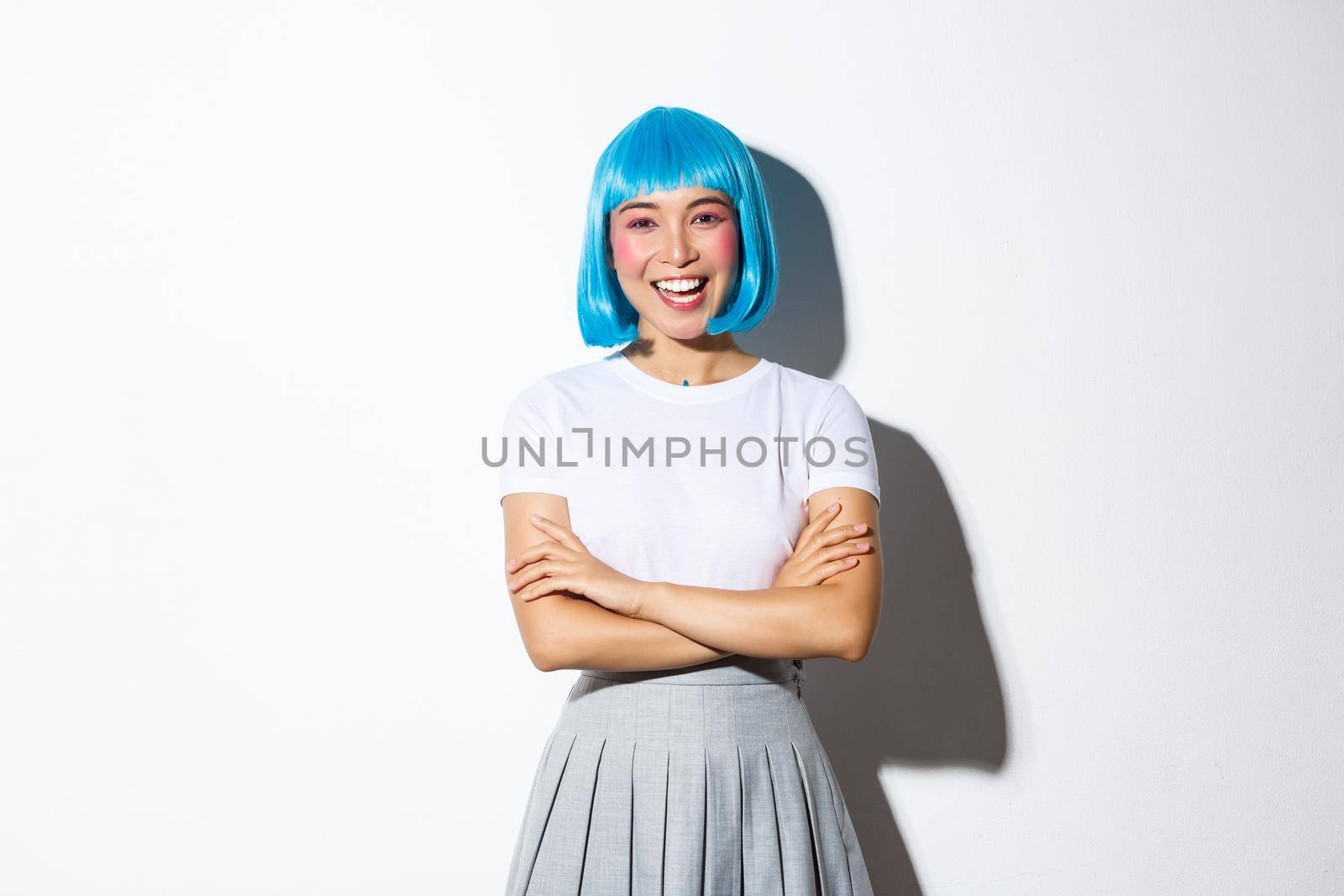 Portrait of confident stylish asian girl in blue short wig, dressed-up for pop party or halloween, cross arms on chest and smiling happy, standing over white background.