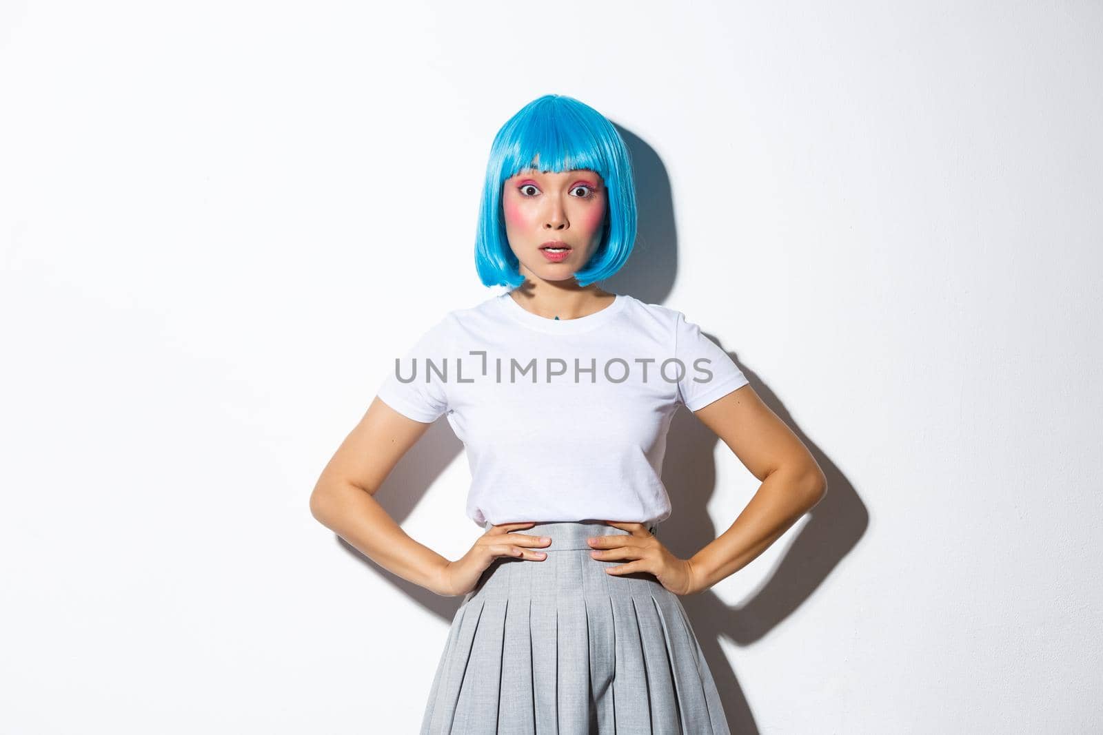 Image of shocked asian girl in blue party wig looking at camera surprised, holding hands on waist in startled pose, standing over white background.