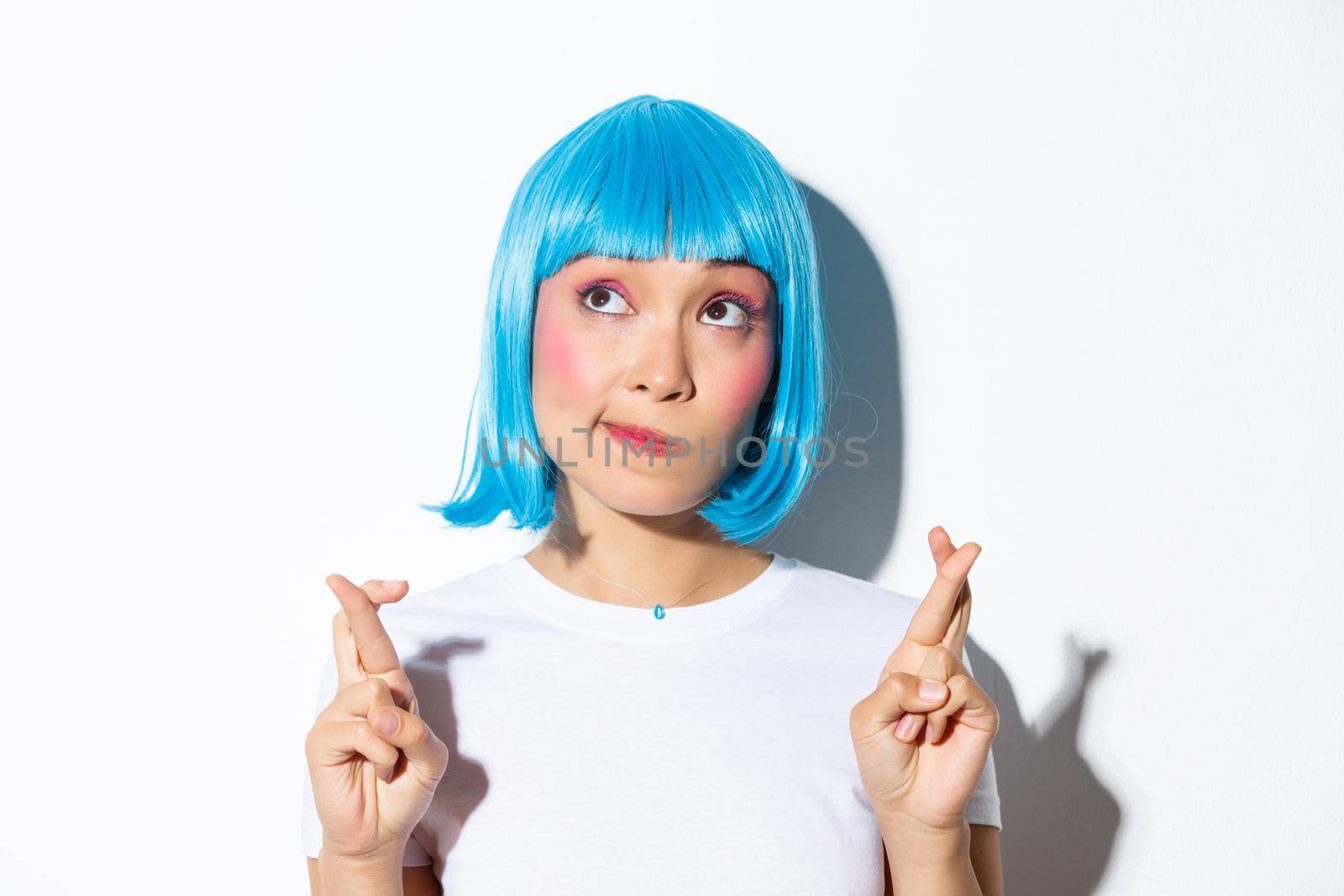 Hopeful beautiful asian girl in blue wig making wish, looking dreamy at upper left corner and cross fingers for good luck, standing over white background.