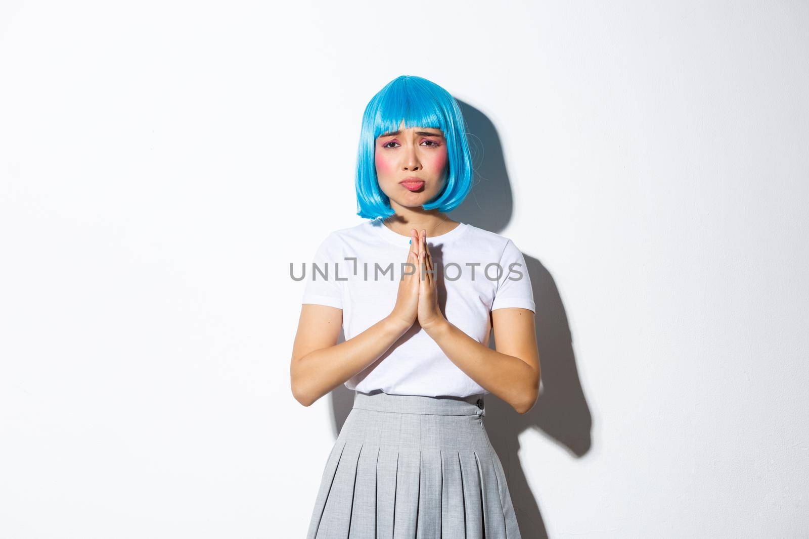 Portrait of sad asian girl in blue wig, begging for help, looking desperate at camera, standing over white background.