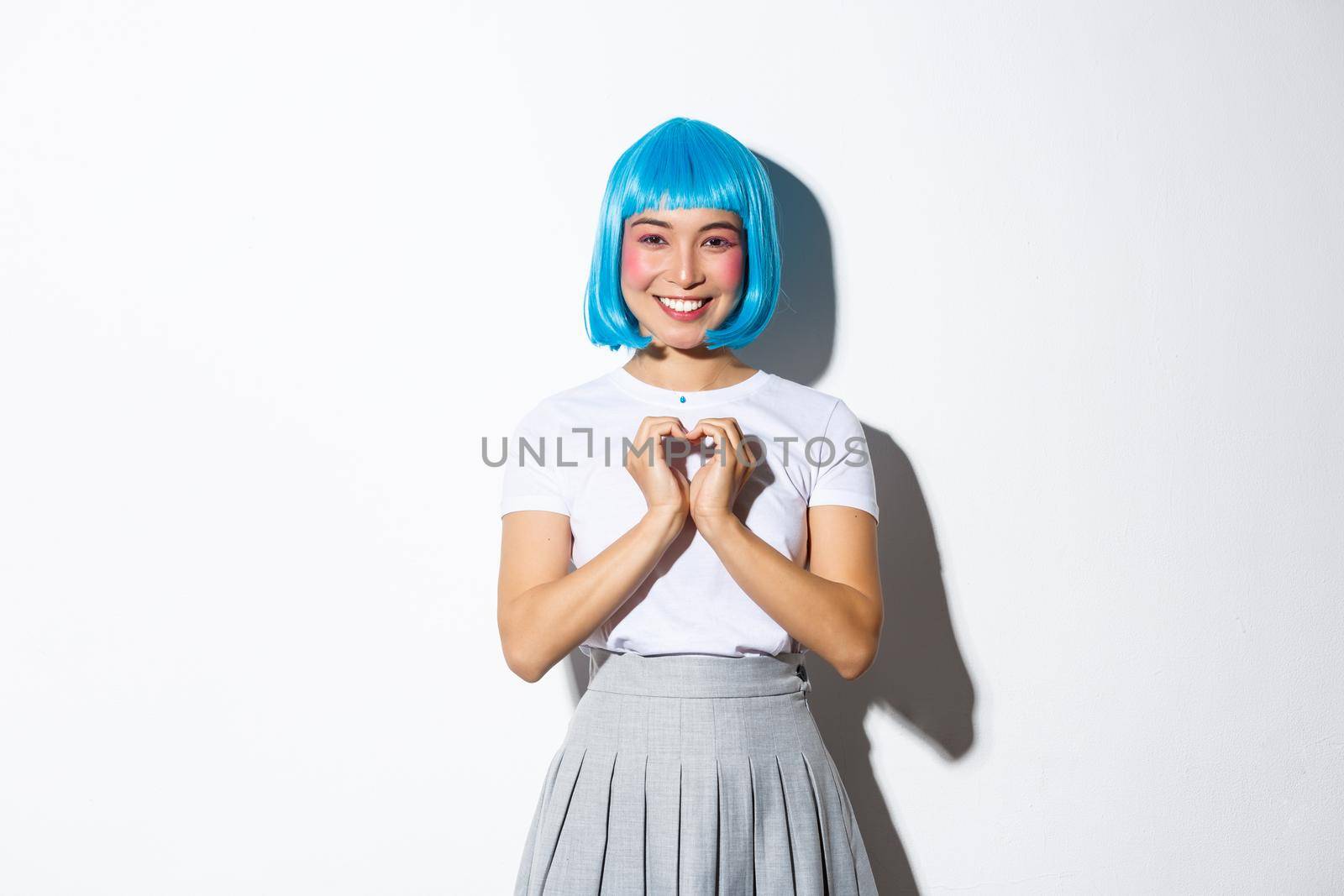 Image of lovely asian girl dressed-up in party costume, wearing wig and showing heart gesture, smiling at camera, standing over white background by Benzoix