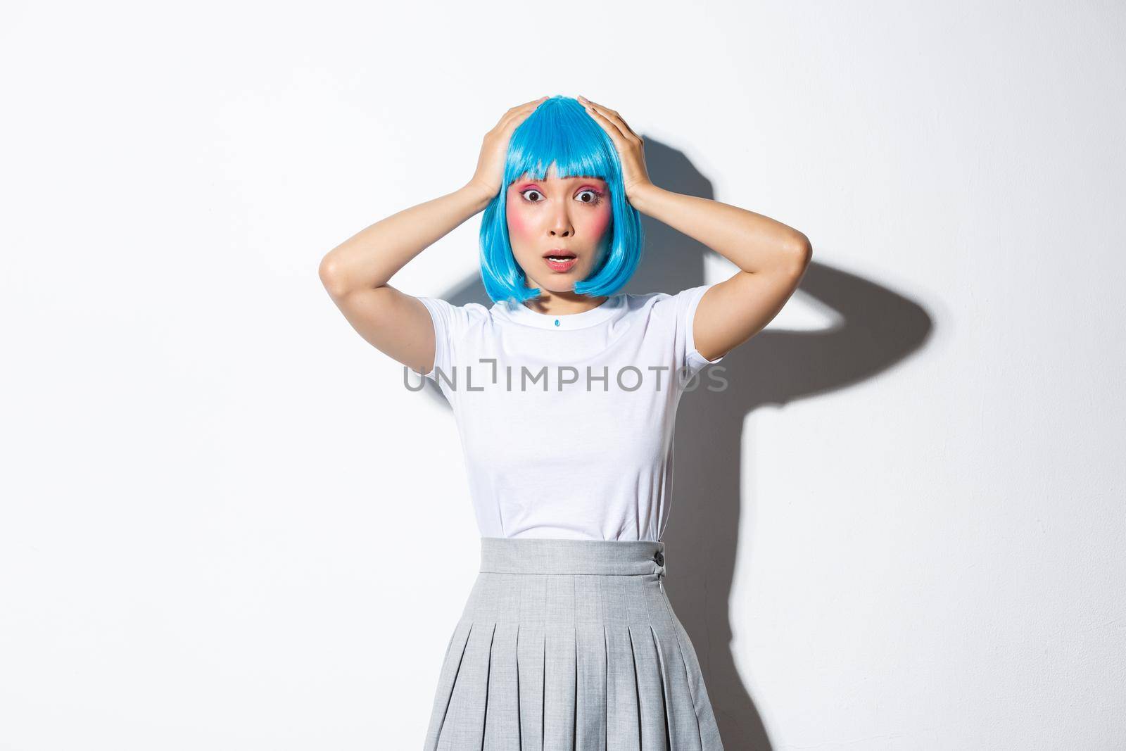 Portrait of shocked and anxious asian girl in blue hair wig, holding hands on head and looking worried, standing over white background in halloween costume.