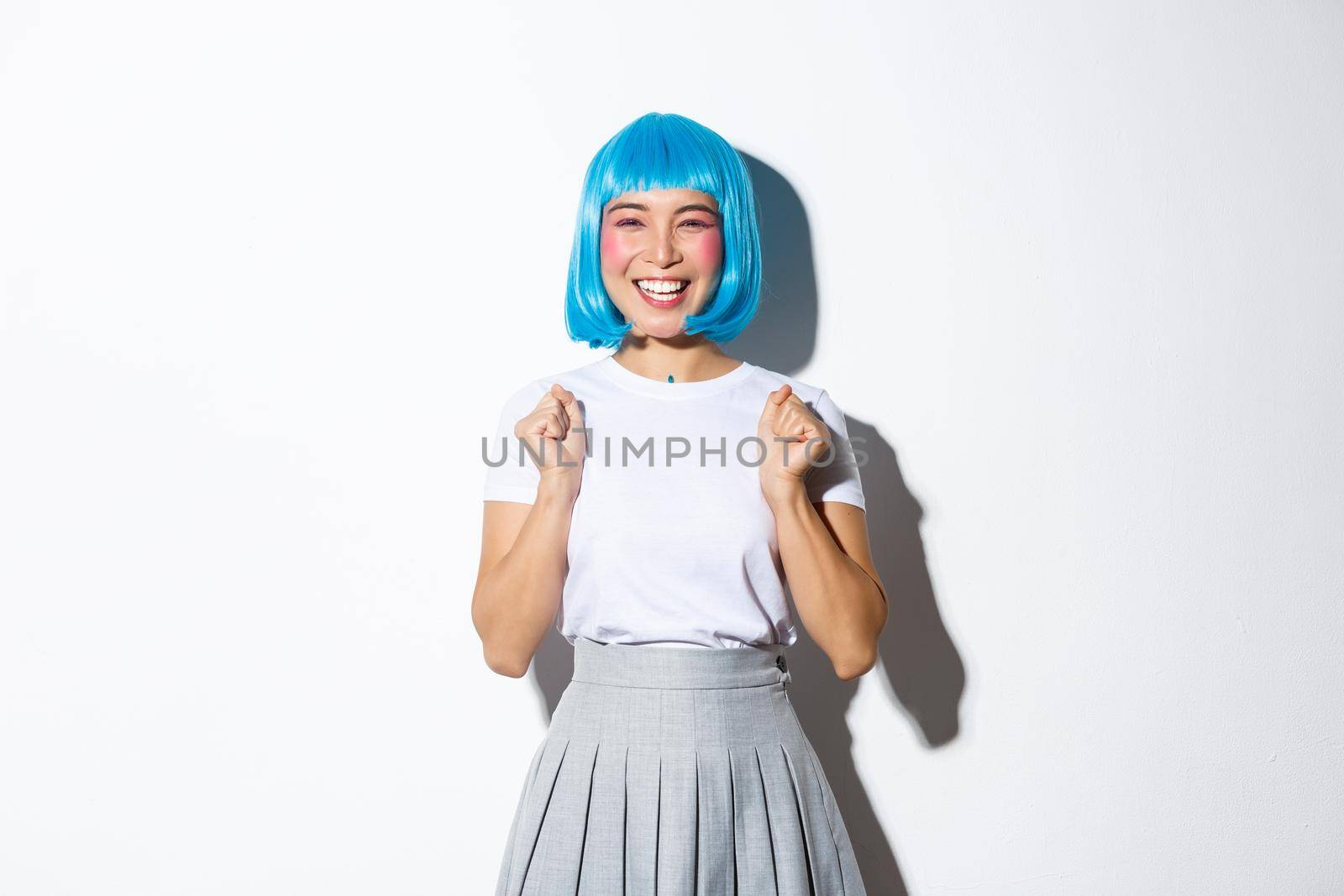 Image of cheerful asian girl feeling like winner, wearing blue wig, celebrating something, smiling and triumphing, standing over white background.