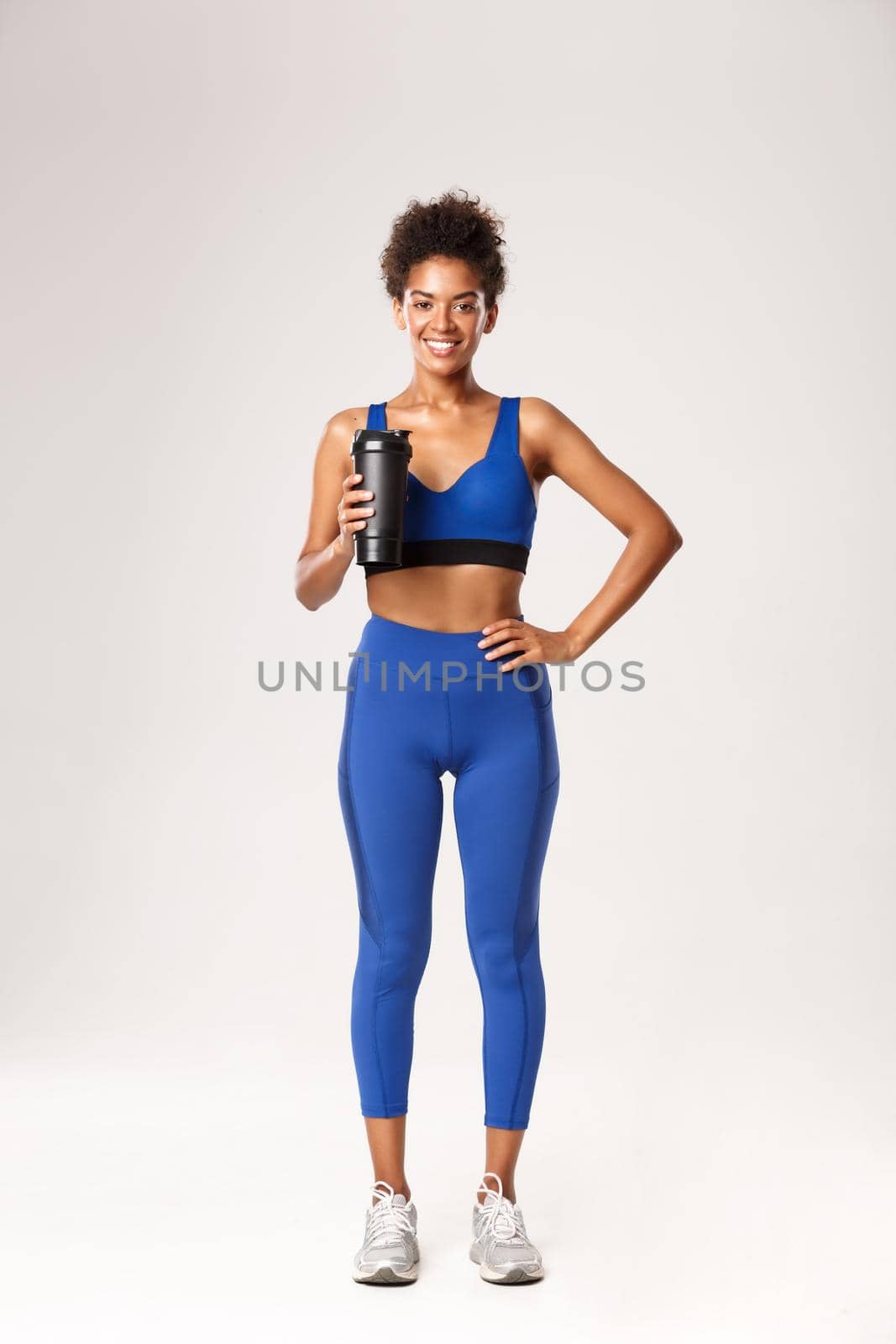 Full length of beautiful african-american fitness woman in blue uniform looking happy, drinking water from bottle, standing against white background.