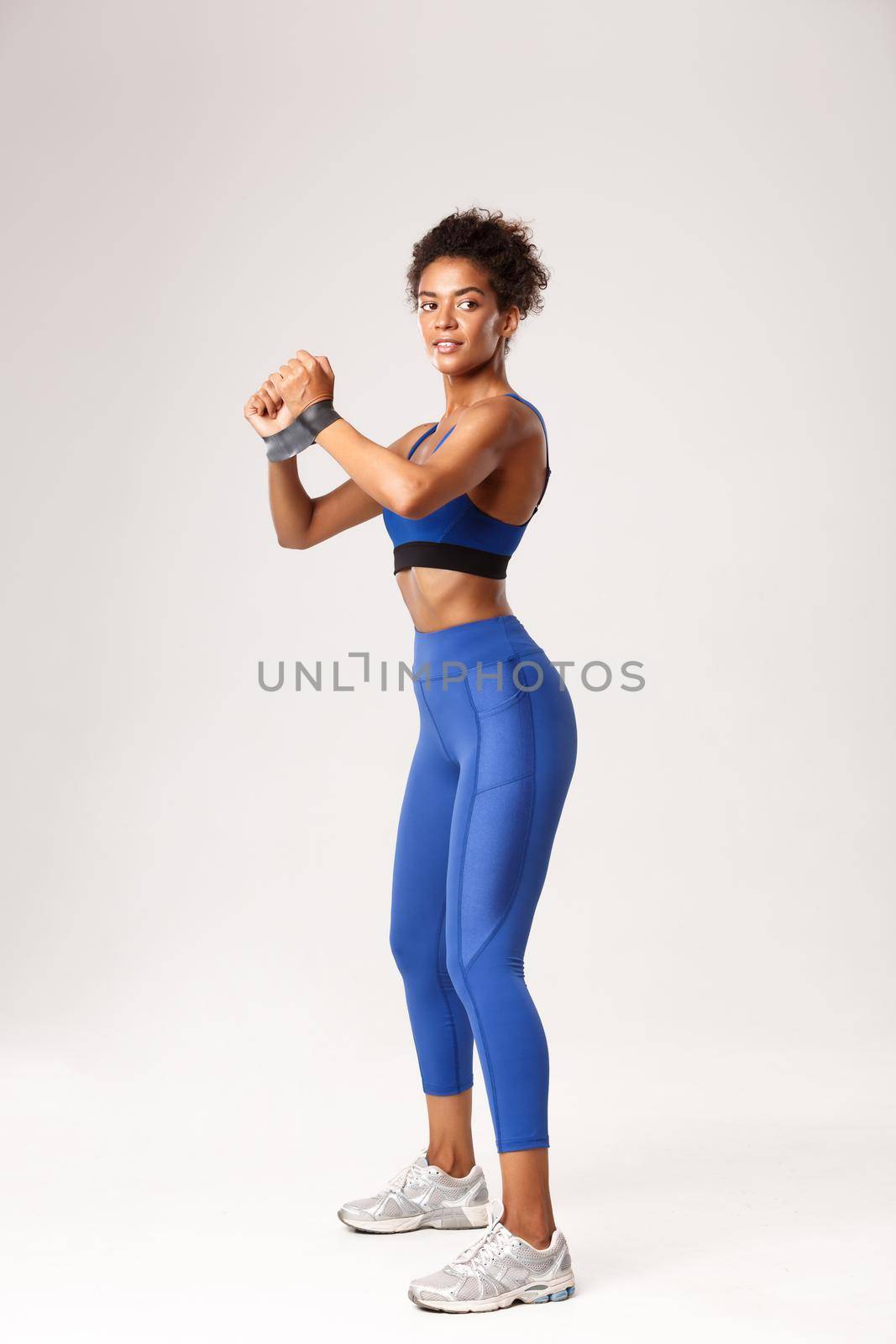Full length profile shot of attractive african-american fitness girl in blue sport outfit, doing exercises with rubber band and looking away, workout against white background.