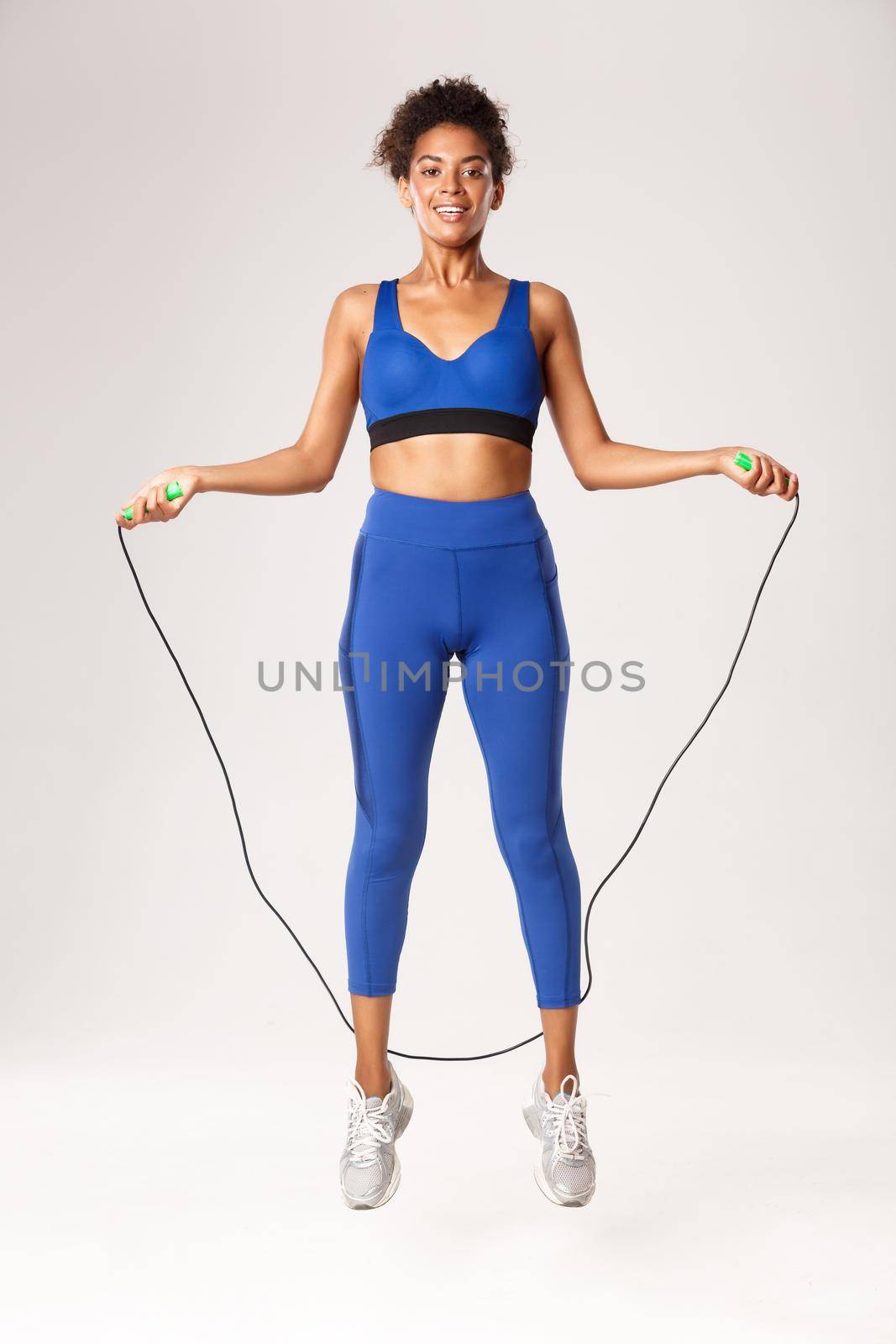 Full length of smiling female athlete in blue sport clothing, jumping with skipping rope and looking happy, workout against white background.