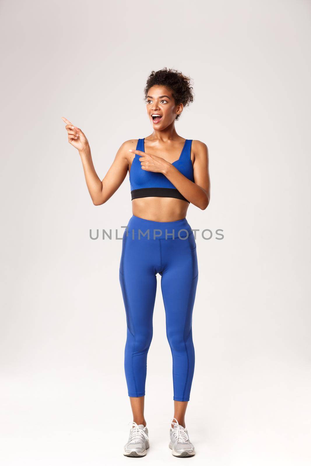 Full length of surprised african-american female athlete in blue sports costume, pointing fingers left and looking with amazement, showing something about workout and gym, white background.