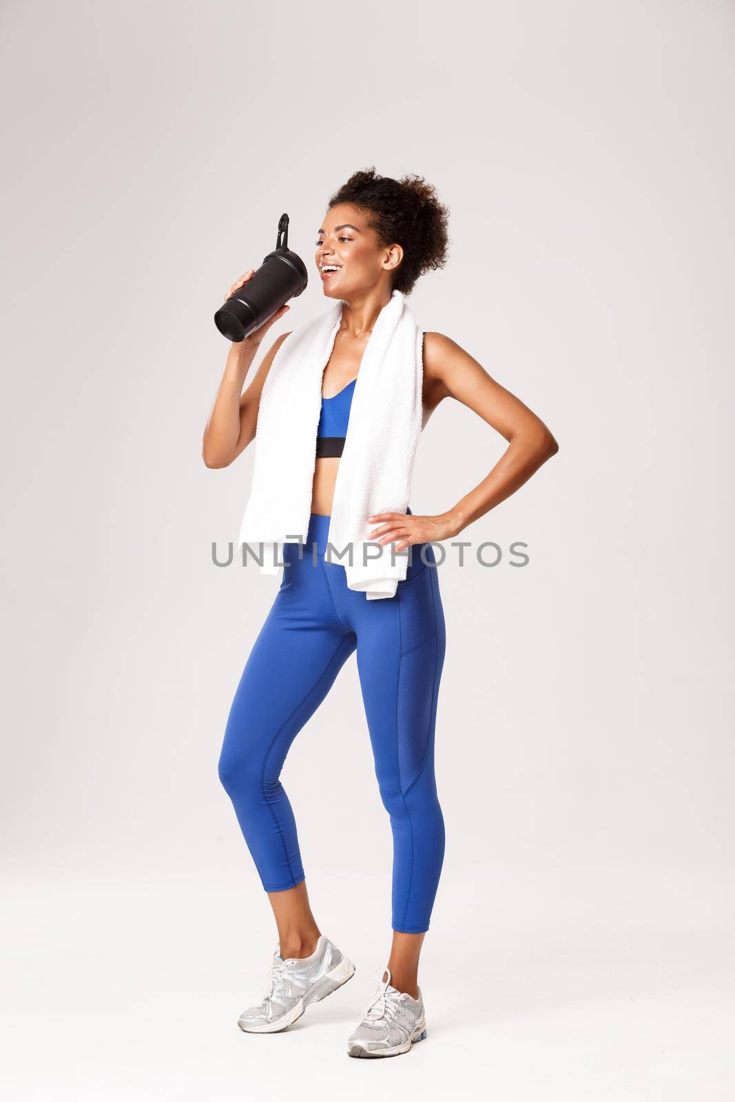 Full length of smiling female athelte in blue sport outfit, drinking protein or water from bottle, standing against white background with towel, finish workout.