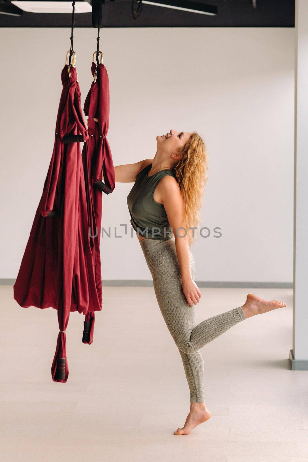 A happy woman does yoga on a suspended burgundy hammock in a bright gym.