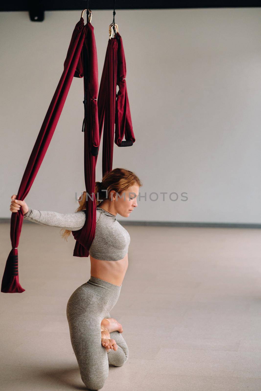A woman does yoga on a suspended burgundy hammock in a bright gym.