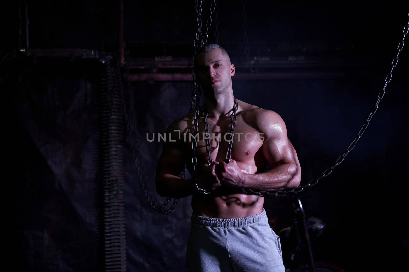 Young shirtless muscular man standing among metal chains, looking seriously at camera, copy space