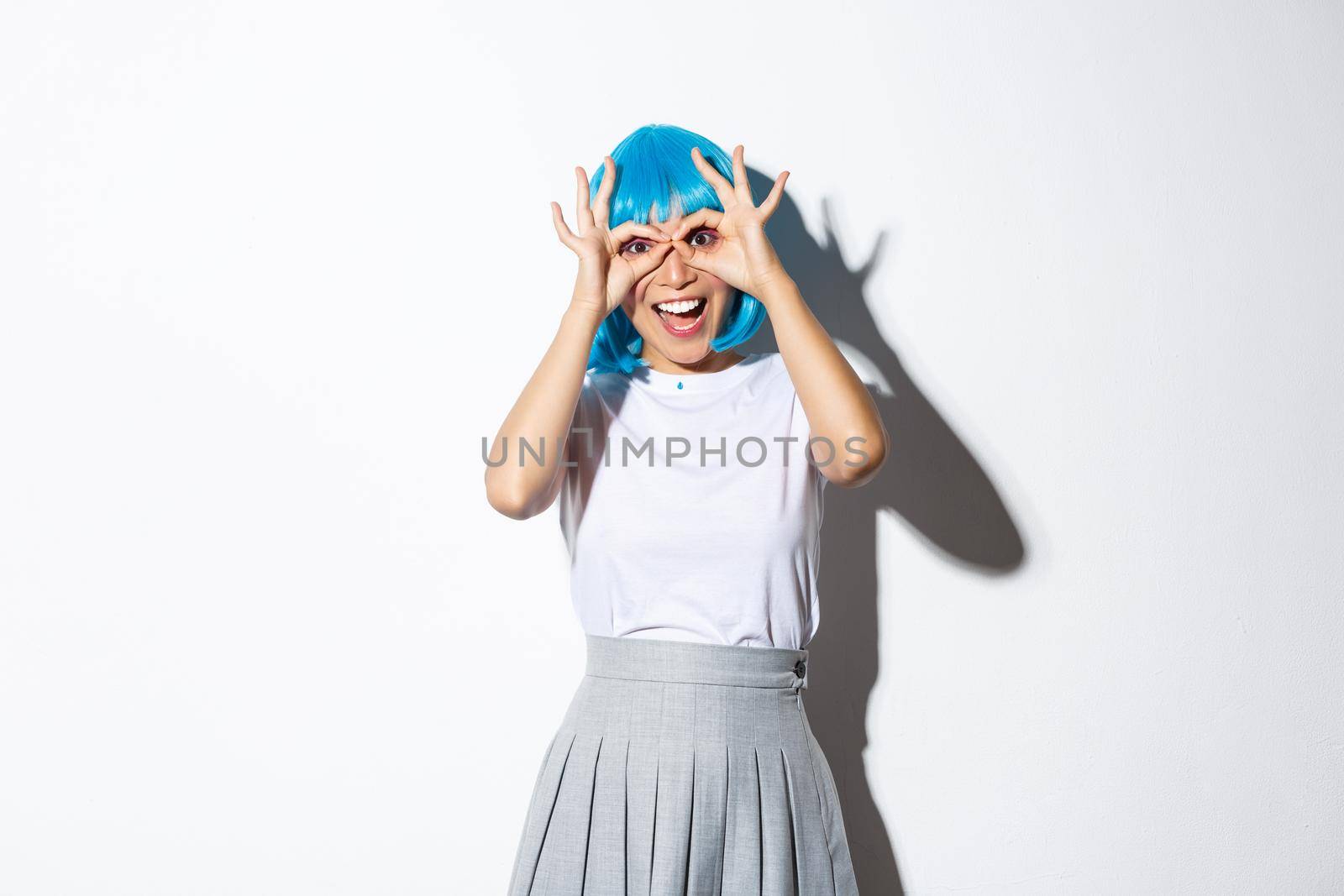 Portrait of cheerful asian girl looking amazed at camera, wearing blue wig and costume for halloween party, standing over white background.