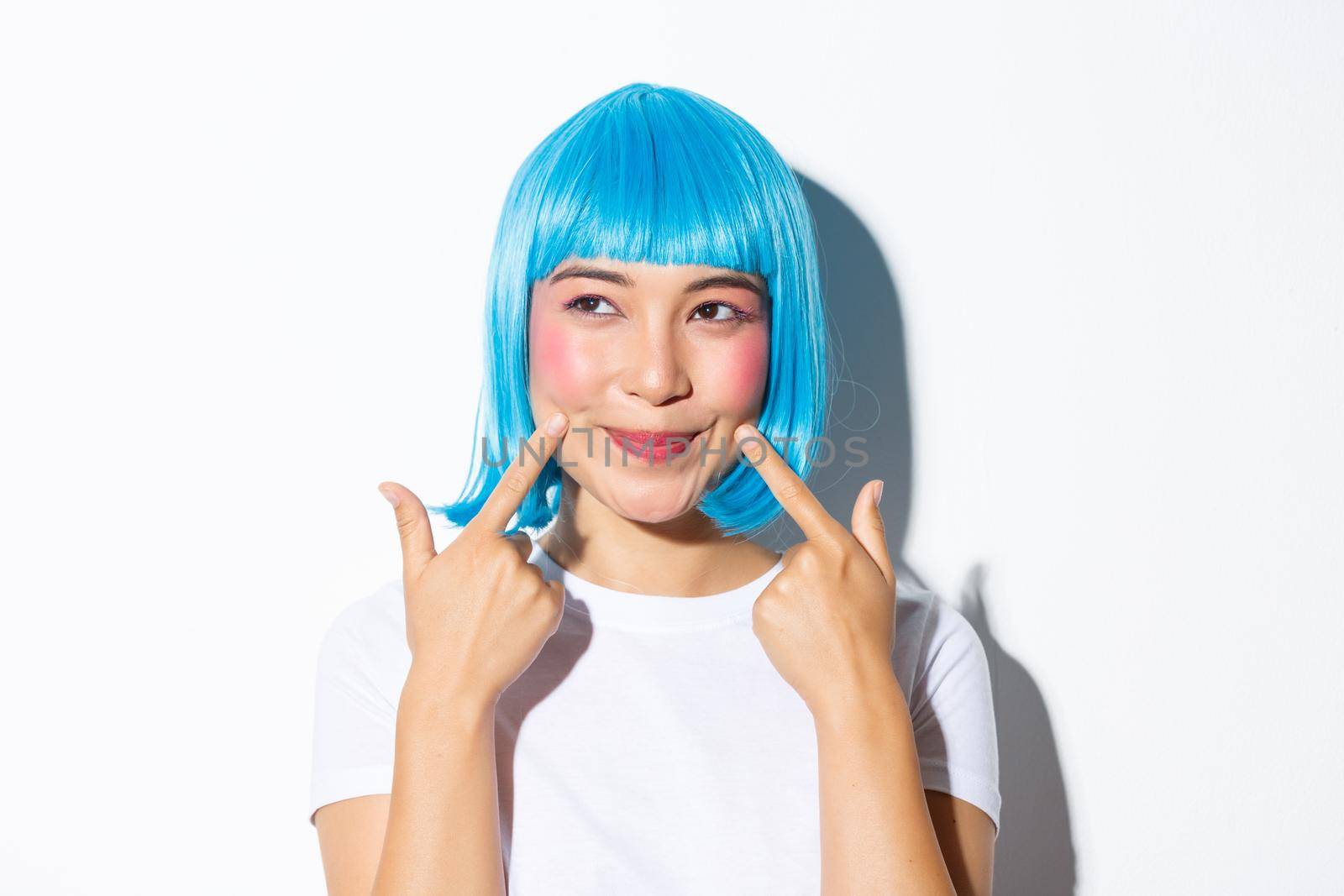 Image of adorable asian girl in blue wig poking her cheeks and looking upper left corner, standing over white background.