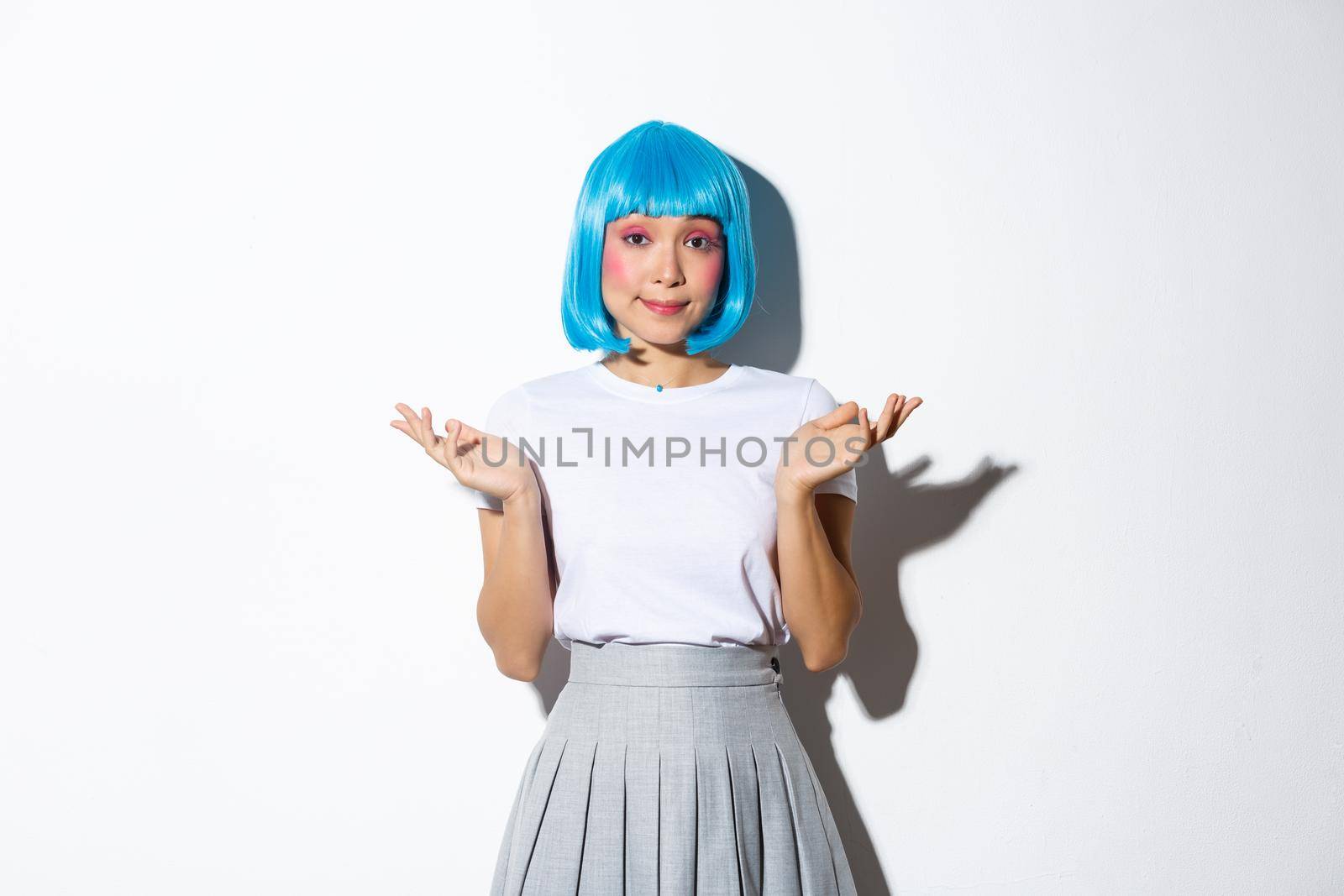 Portrait of clueless young asian party girl in blue wig shrugging, looking indecisive at camera, standing over white background.