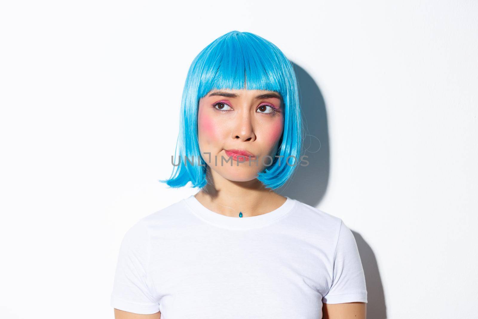 Close-up of doubtful frowning asian girl in blue wig, looking with disbelief at upper left corner, standing over white background.