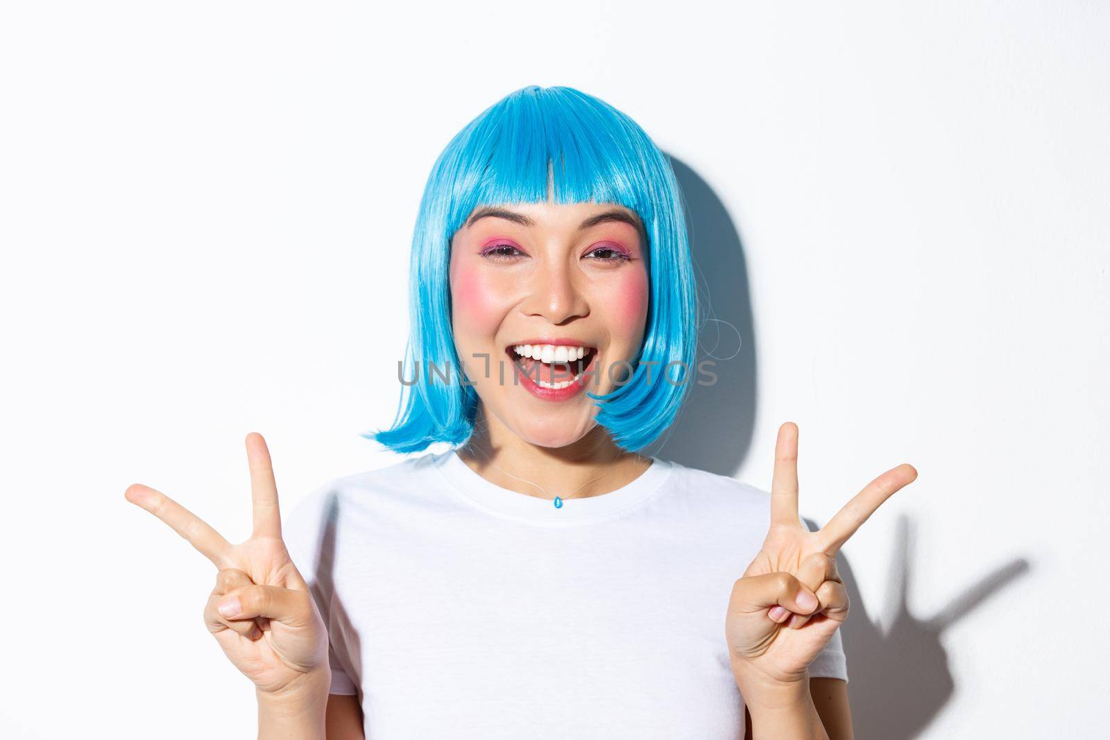 Close-up of beautiful excited asian girl showing peace gesture and smiling, celebrating holiday, wearing blue wig for halloween party.