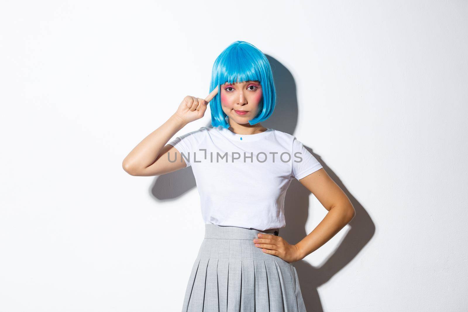 Image of asian girl in blue wig looking skeptical, pointing finger at head, standing over white background.