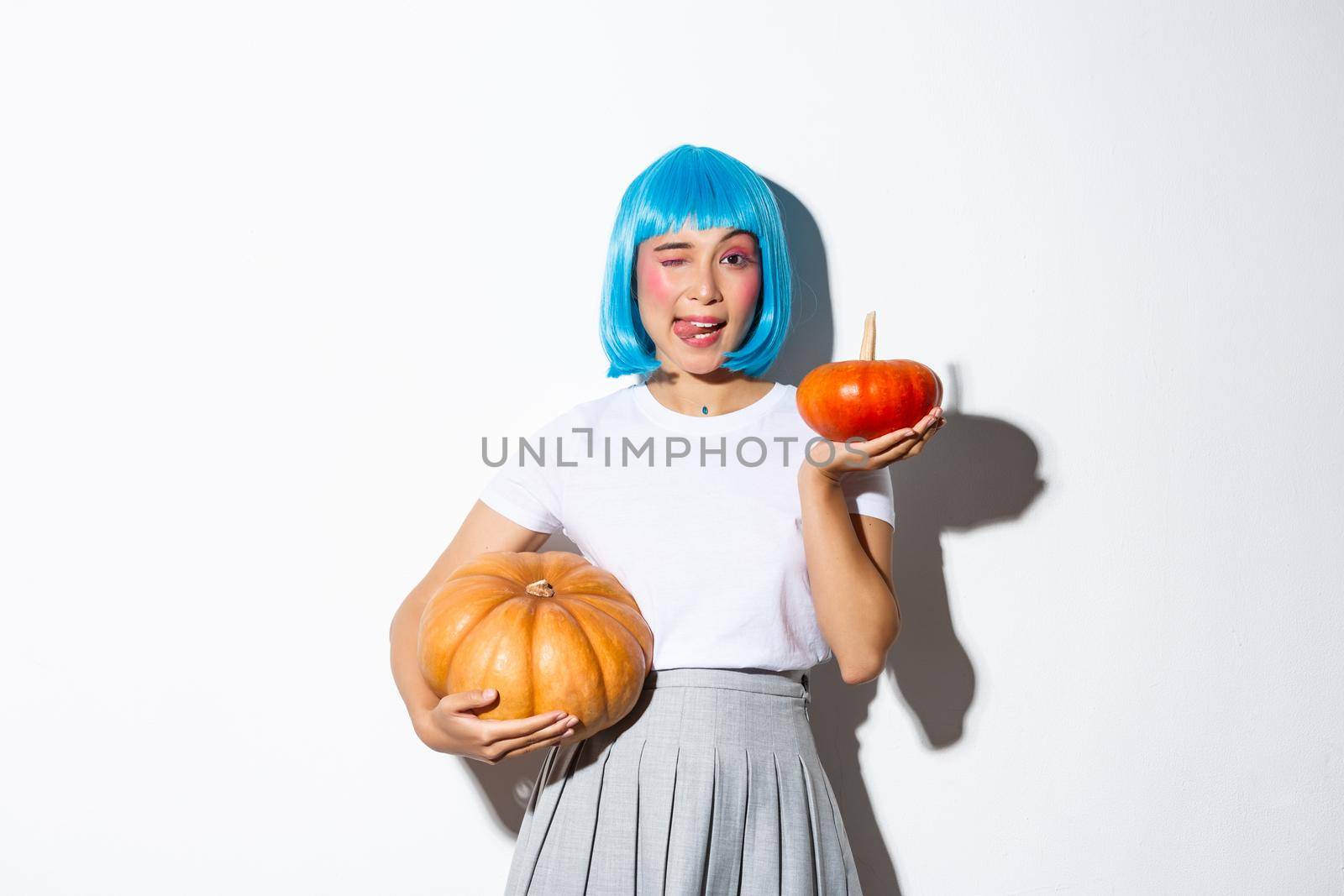 Cute asian woman in blue wig, showing tongue and winking coquettish, holding two pumpkins, celebrating halloween.