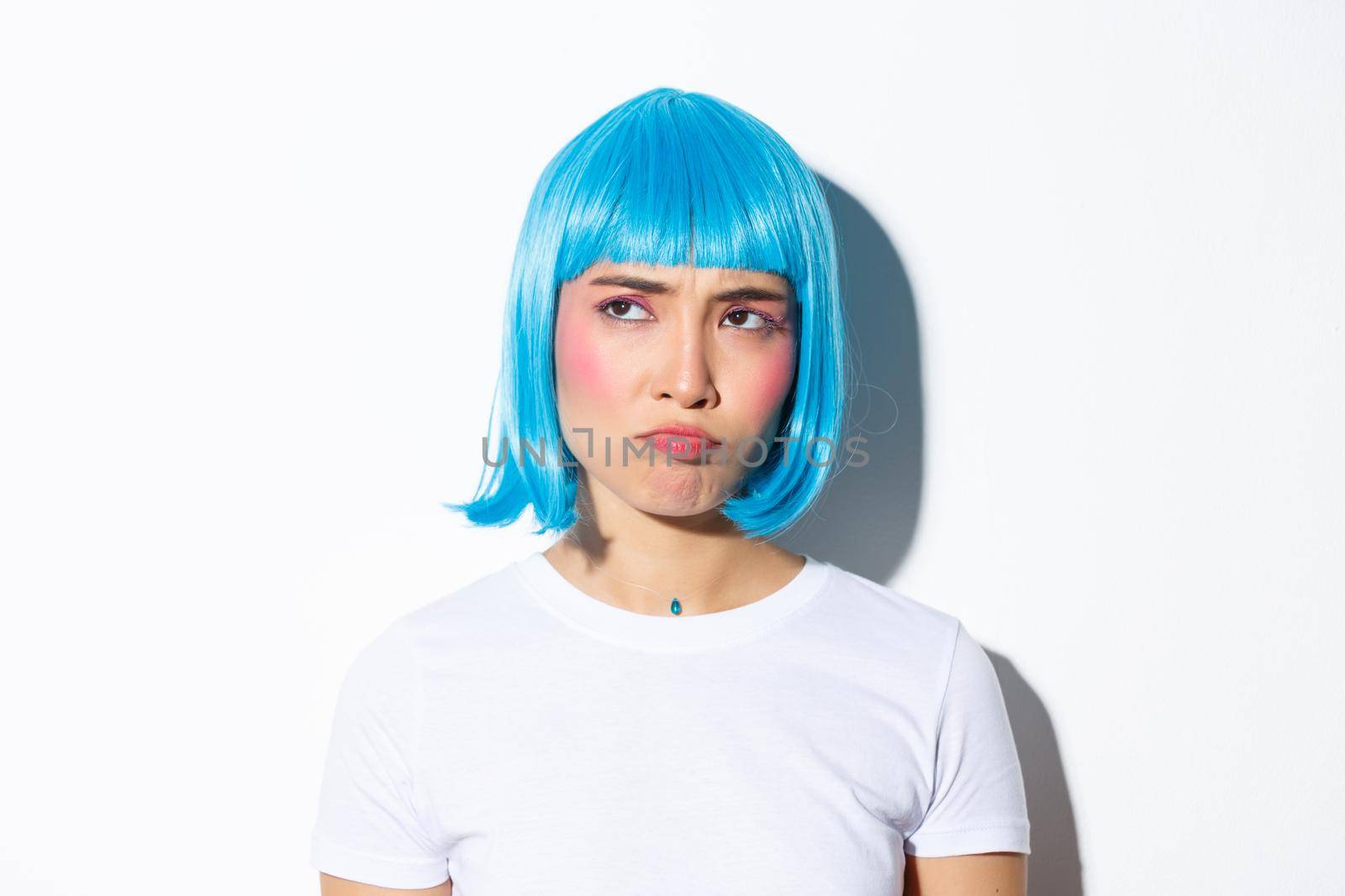 Image of cute asian girl in blue wig looking disappointed or jealous, sulking while stare at upper left corner, standing against white background.