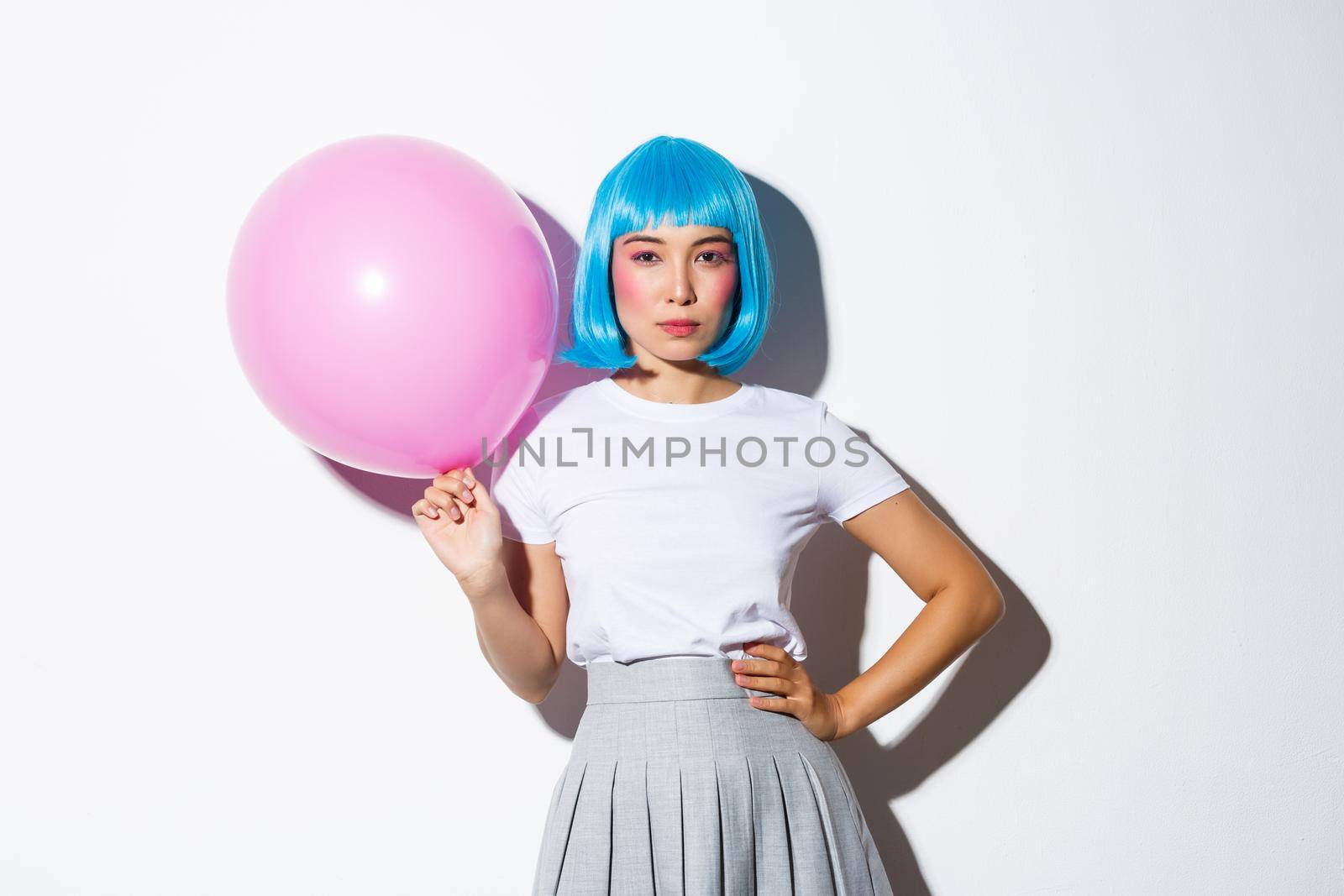 Image of sassy asian woman in blue wig, looking determined, holding pink balloon, standing over white background.