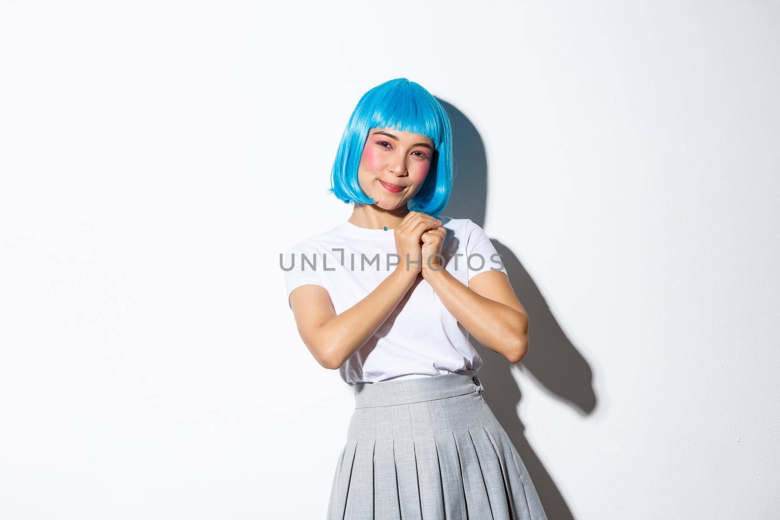 Portrait of cute beautiful asian girl in blue wig, clasp hands together and looking with admiration, standing over white background.