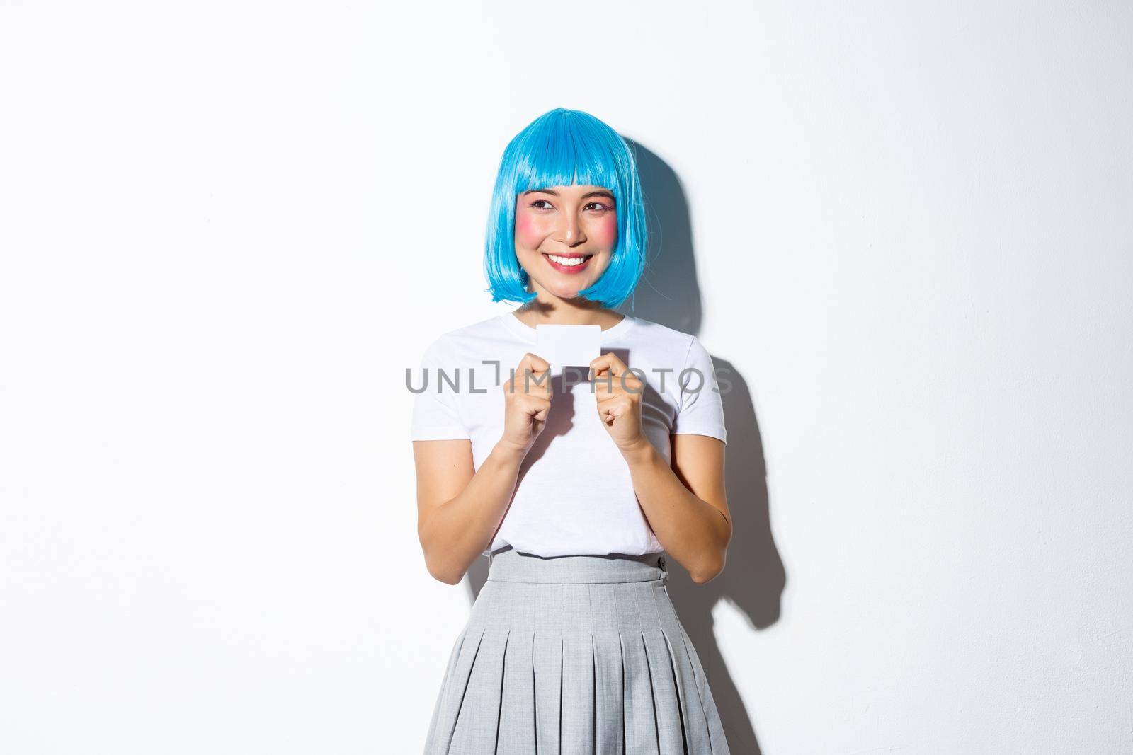 Image of happy asian girl in blue wig and halloween costume, looking upper left corner thoughtful, smiling and showing credit card, standing over white background.