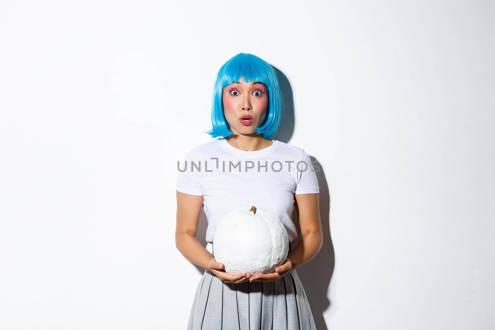 Image of asian girl in blue wig looking surprised and gasping, holding white pumpkin, standing over white background.