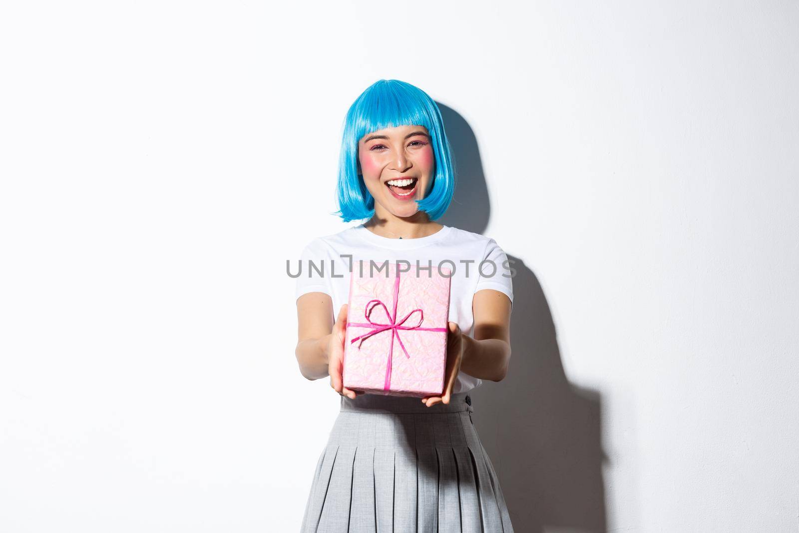 Portrait of cute happy asian girl in blue party wig giving you gift, standing over white background.