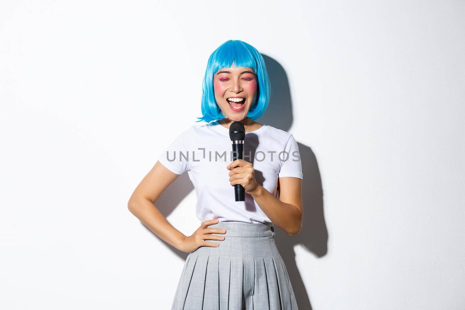 Beautiful asian female singing karaoke and smiling, wearing blue wig for halloween party, standing over white background.