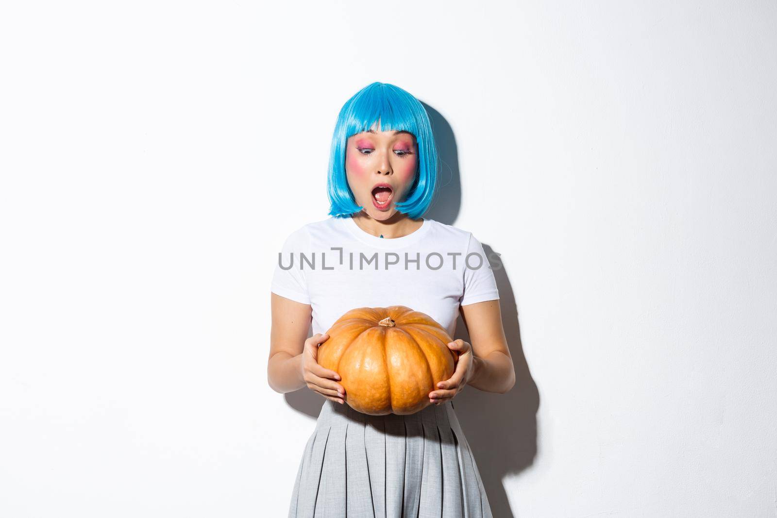 Image of amazed asian girl in blue wig drop jaw, looking at large pumpkin astonished, standing over white background.