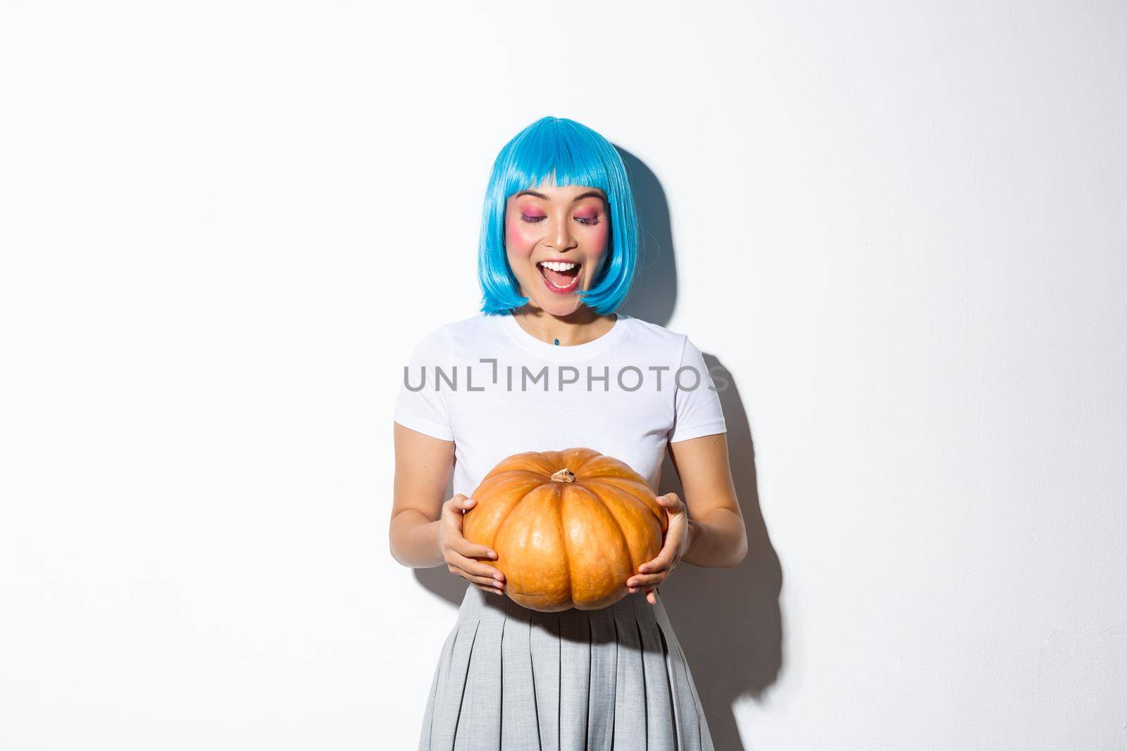 Beautiful smiling asian woman celebrating halloween in blue wig, looking at pumpkin with happy expression, standing over white background.
