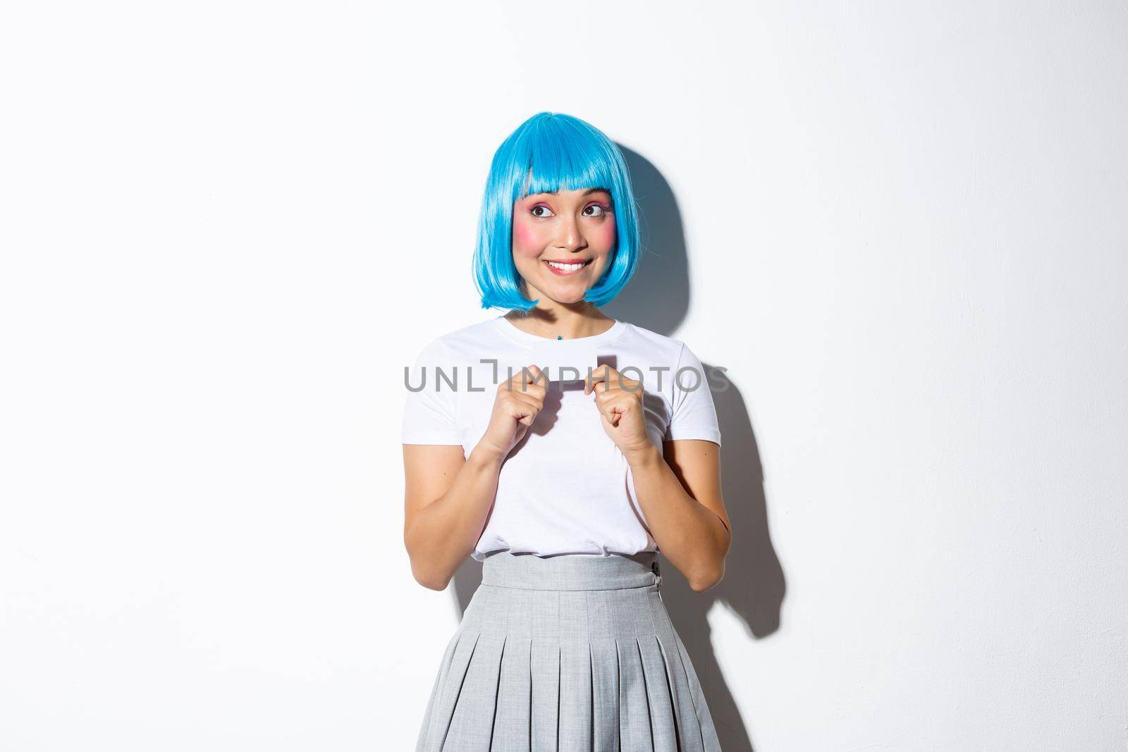 Portrait of silly smiling girl dreaming of shopping, looking upper left corner tempted and holding credit card, wearing halloween costume with blue short wig.