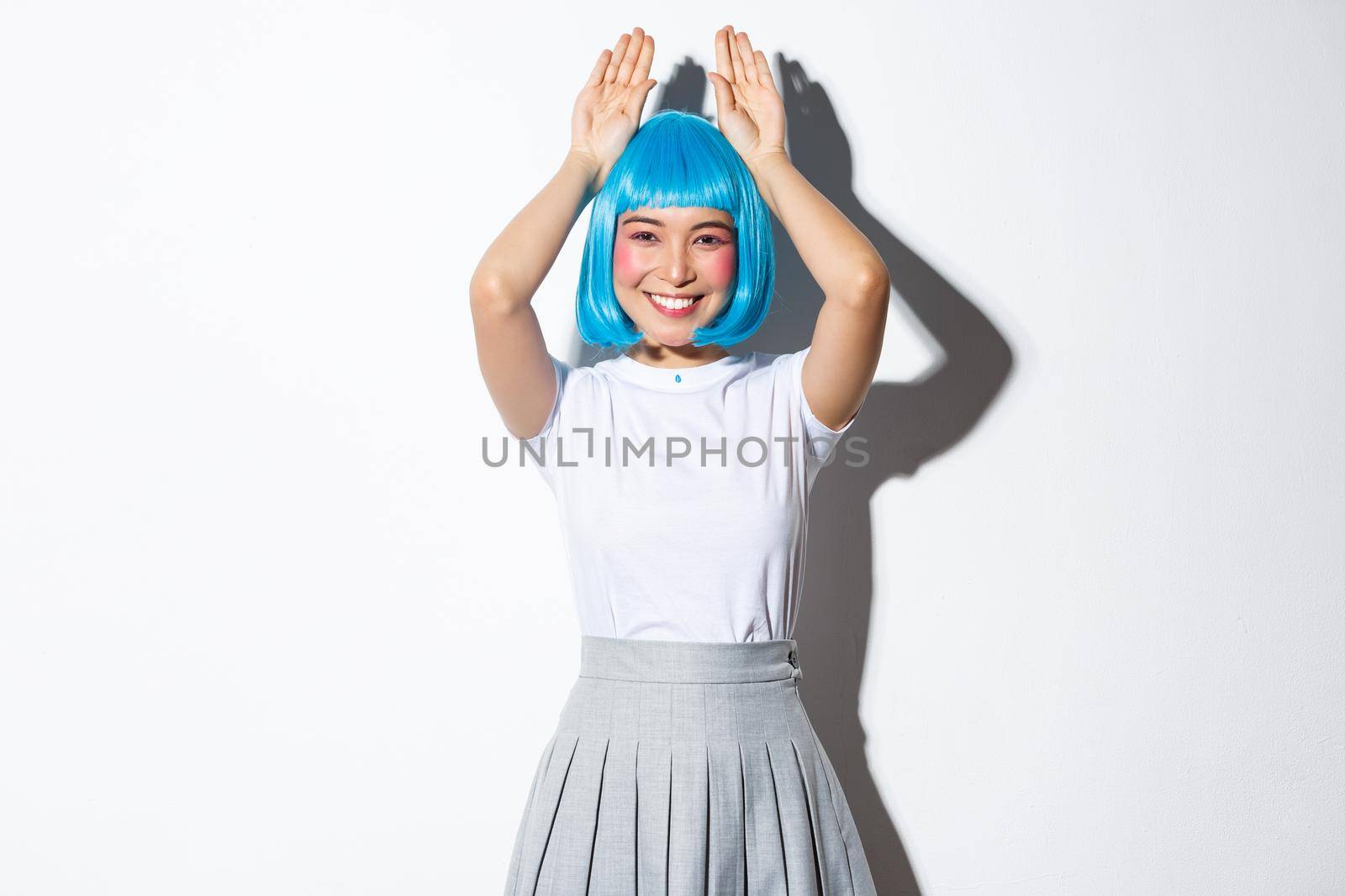 Image of beautiful asian girl smiling and making bunny ears gesture, wearing blue wig for halloween party, standing over white background.
