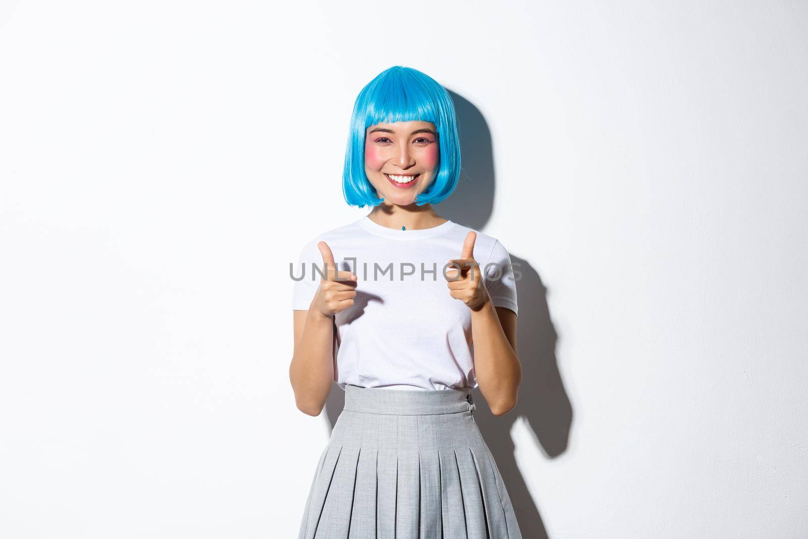 Cheeky japanese girl in blue party wig pointing fingers at camera and smiling, celebrating halloween, standing over white background by Benzoix