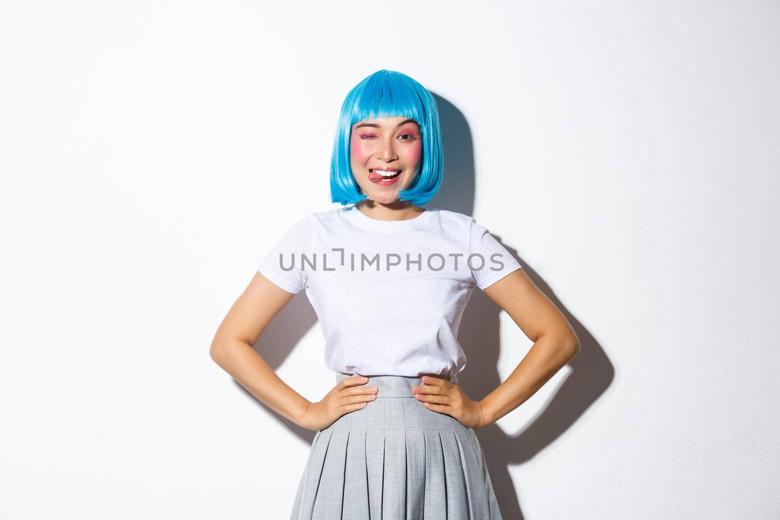 Portrait of beautiful sassy asian girl in blue party wig, looking happy and winking, standing over white background.
