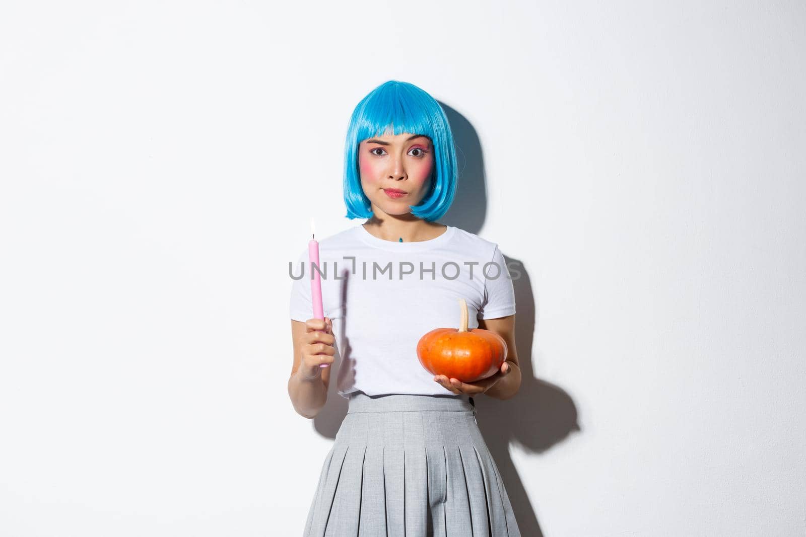 Concept of halloween. Image of attractive skeptical asian girl in blue wig, holding candle and pumpkin, looking serious at camera.