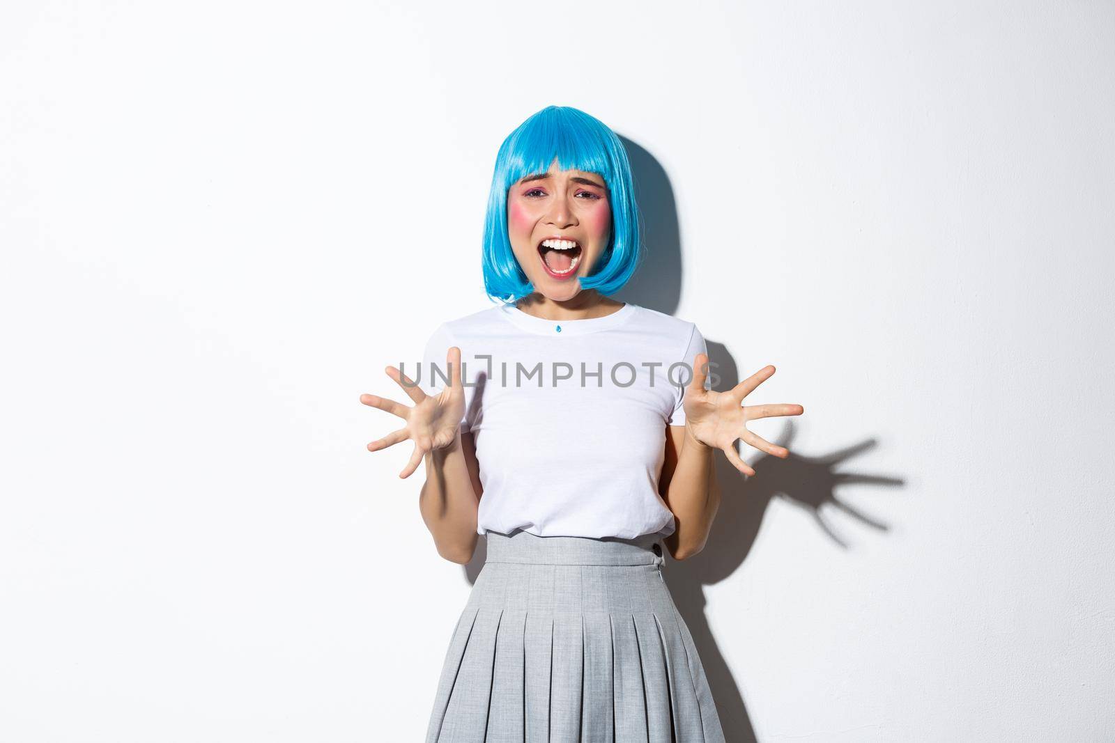 Portrait of asian girl wearing blue wig as a costume for halloween, screaming from fear, looking horrified at camera, standing over white background.