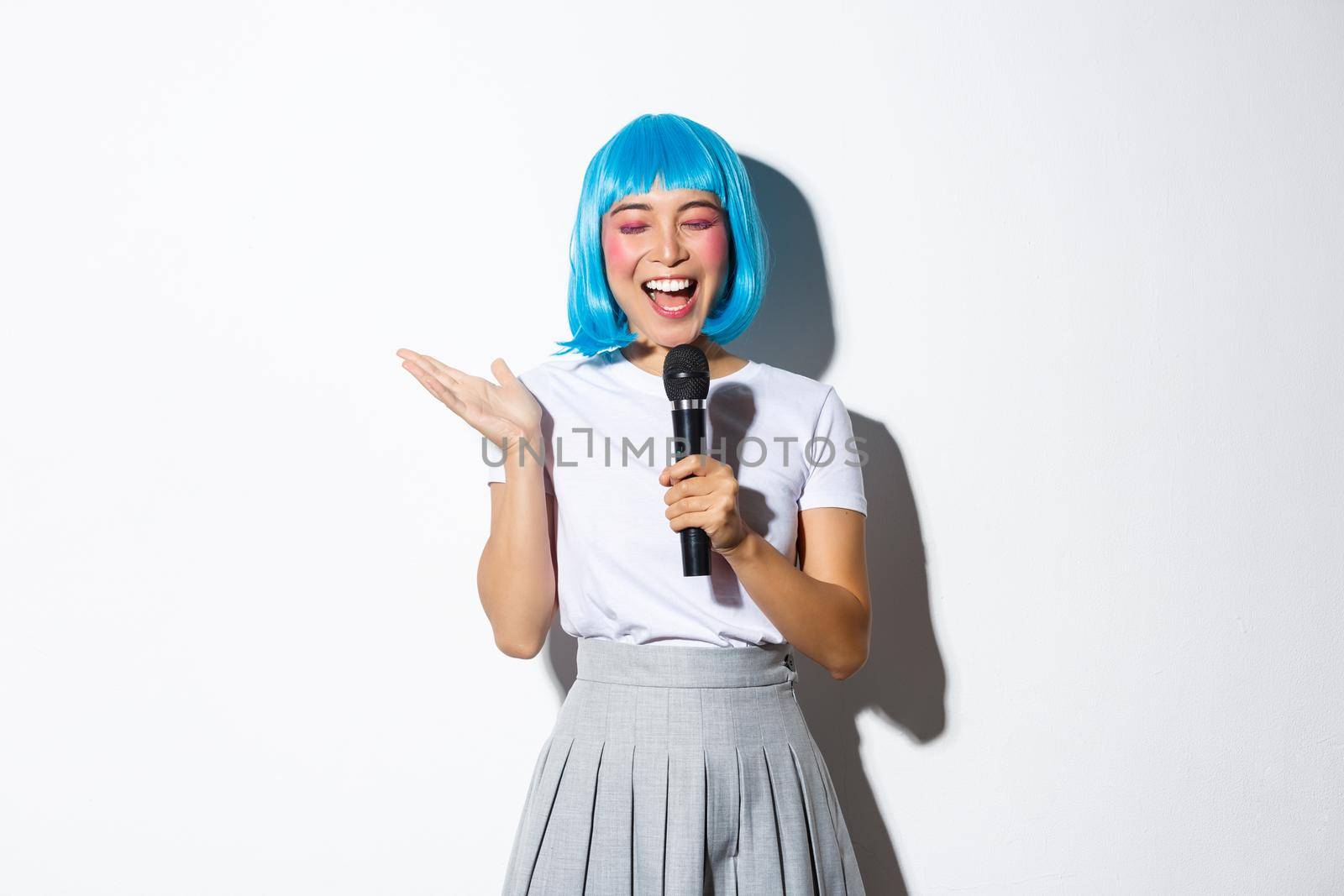 Cheerful cute asian girl dressed up as anime character for halloween party, wearing blue wig and schoolgirl costume, holding microphone and singing karaoke, standing over white background.