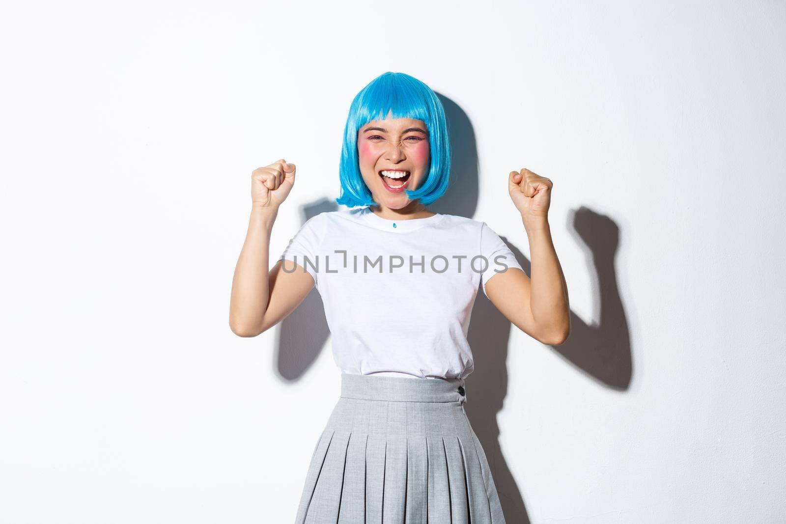 Portrait of successful happy girl in blue wig looking like winner, raising hands up and laughing, standing over white background.
