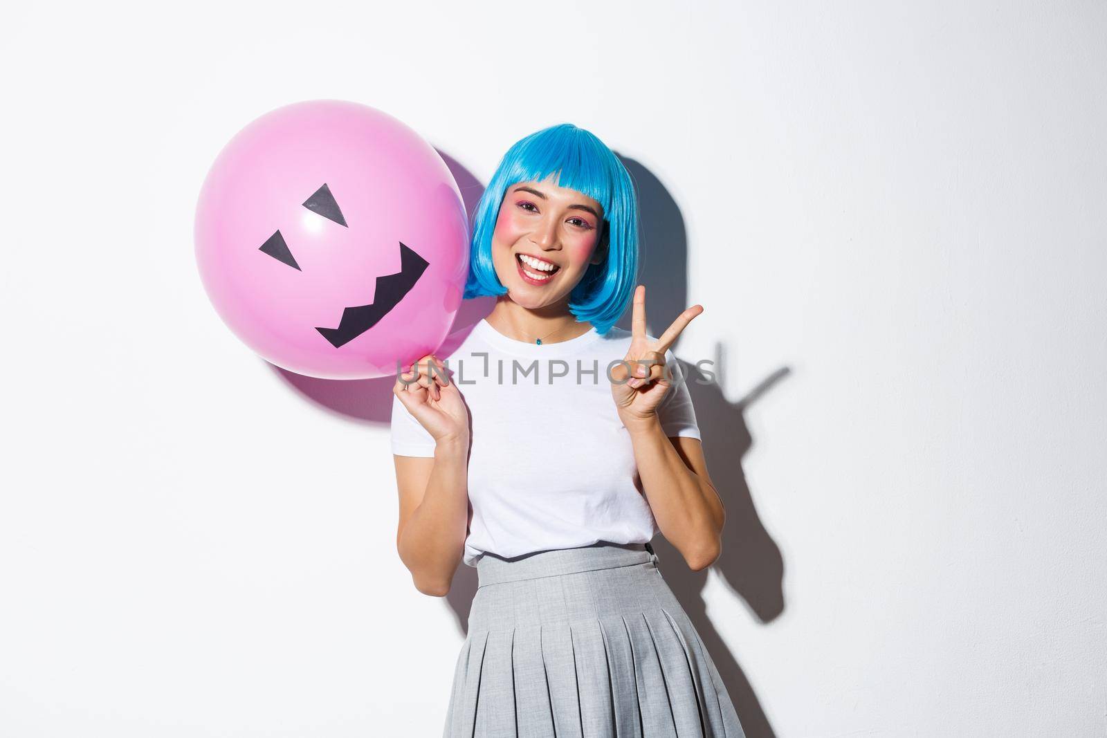 Image of happy asian girl celebrating halloween in blue anime wig, holding pink balloon with scary face and showing peace gesture by Benzoix