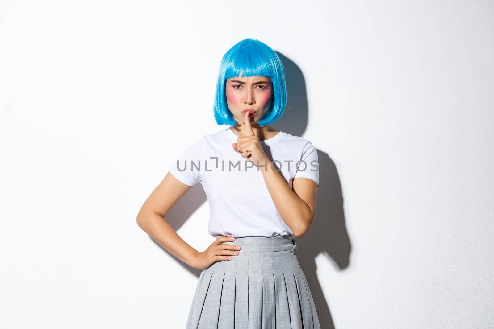 Angry disappointed asian girl scolding someone, shushing at camera and frowning mad, wearing blue wig for halloween party, standing over white background.