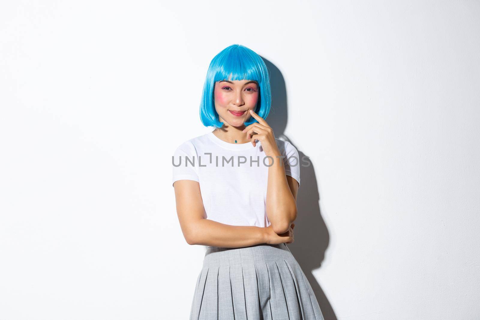 Image of cunning smiling asian girl in blue party wig, looking devious at camera, have an idea, standing over white background.