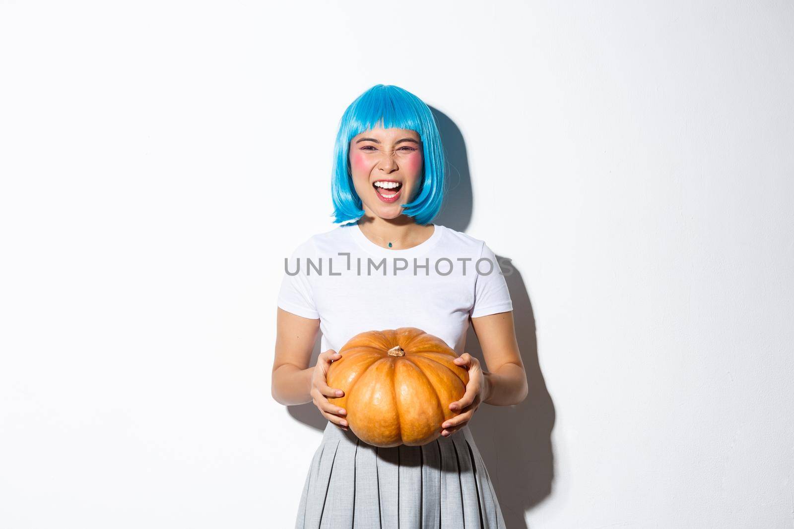 Sassy attractive girl partying, celebrating halloween in blue wig and holding pumpkin, standing over white background.