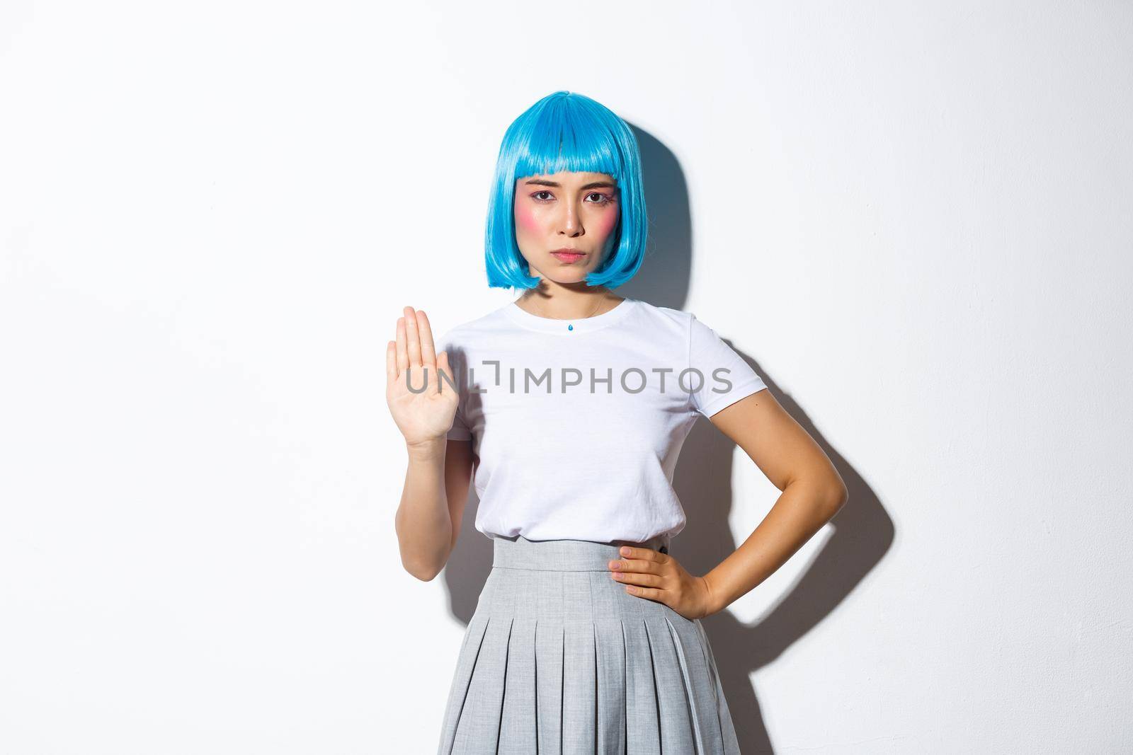 Portrait of serious and confident asian woman in blue wig showing stop gesture, disapprove or disagree with someone, standing over white background.