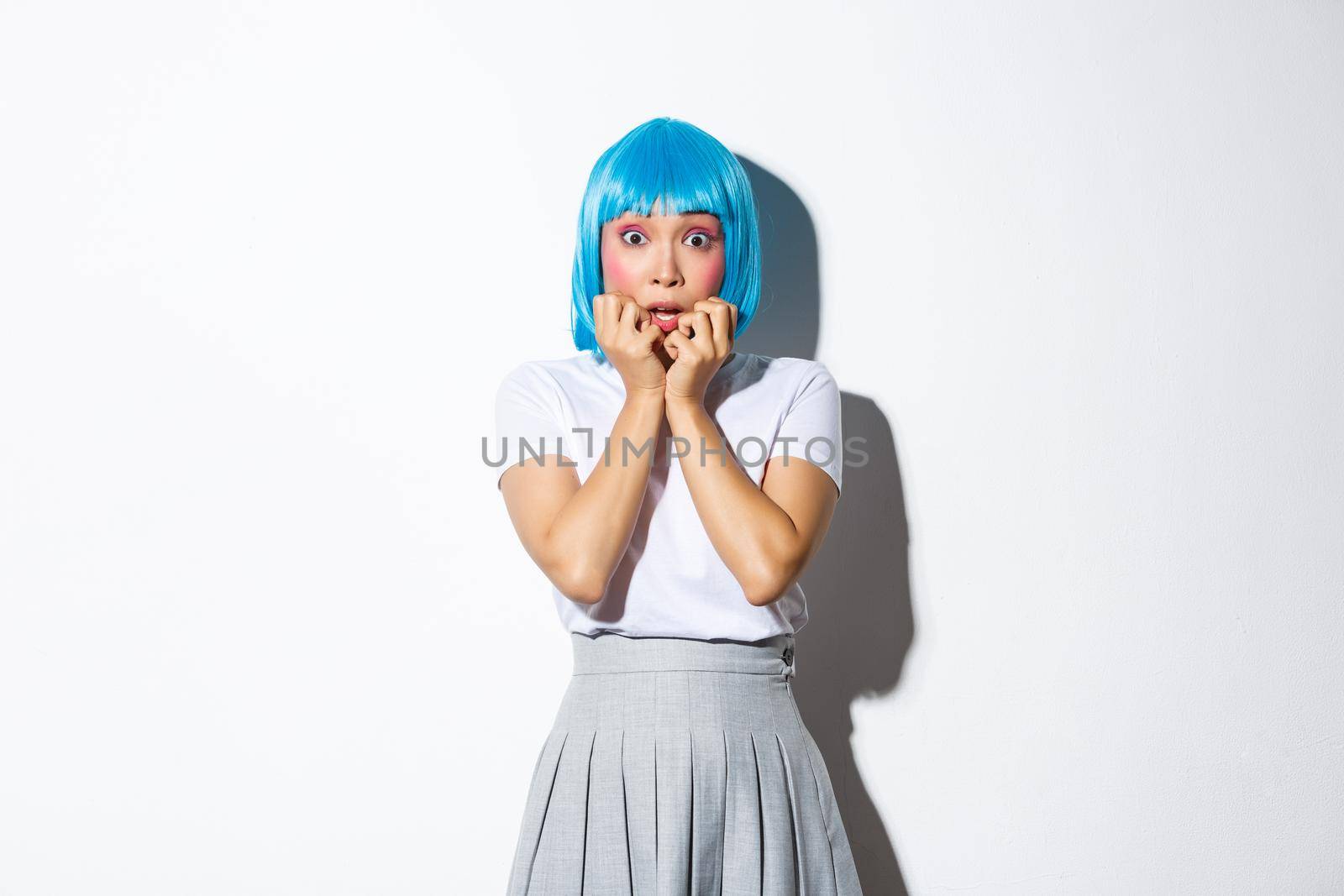 Portrait of scared asian woman gasping afraid, stare at camera ambushed, wearing blue party wig and schoolgirl uniform for halloween, standing over white background.