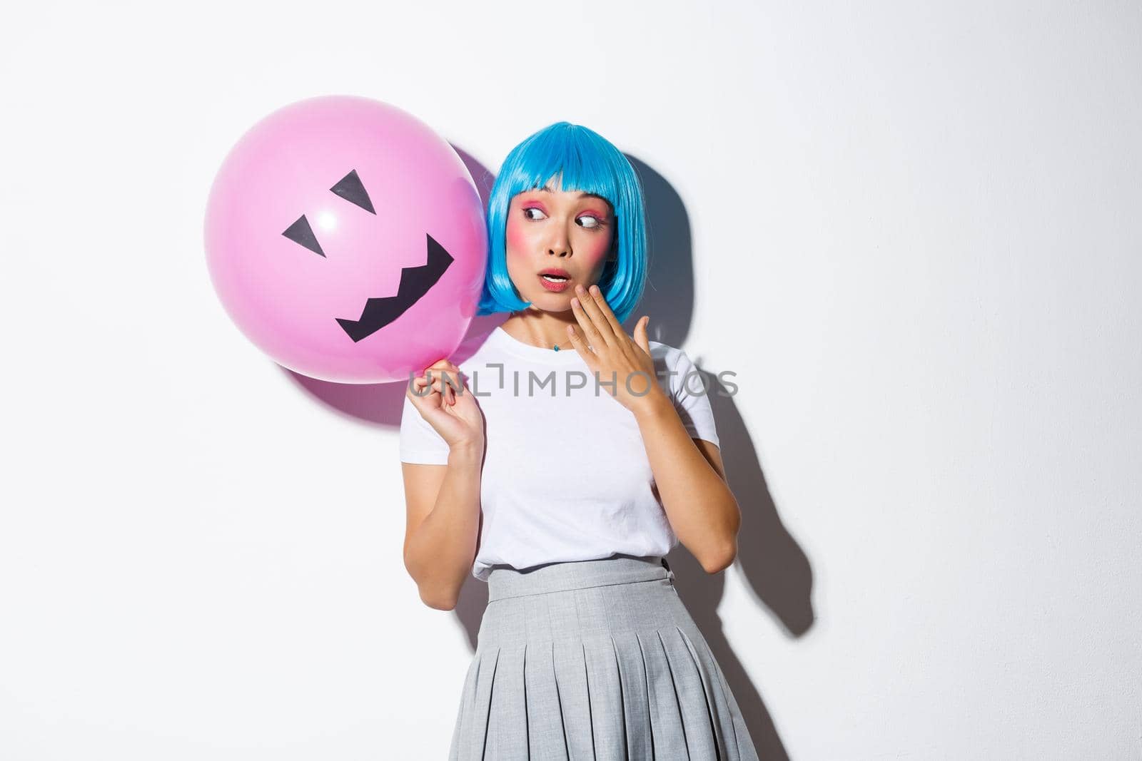 Cute and silly asian girl in blue wig, celebrating halloween, looking surprised at balloon with scary face.