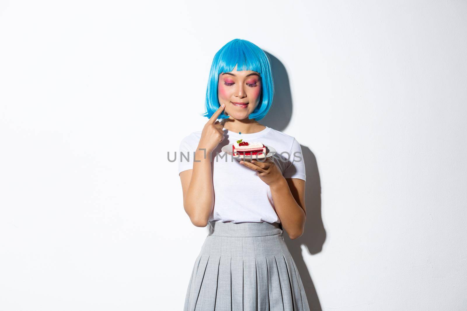 Image of coquettish fashion girl in blue wig looking with temptation at cake, standing over white background.